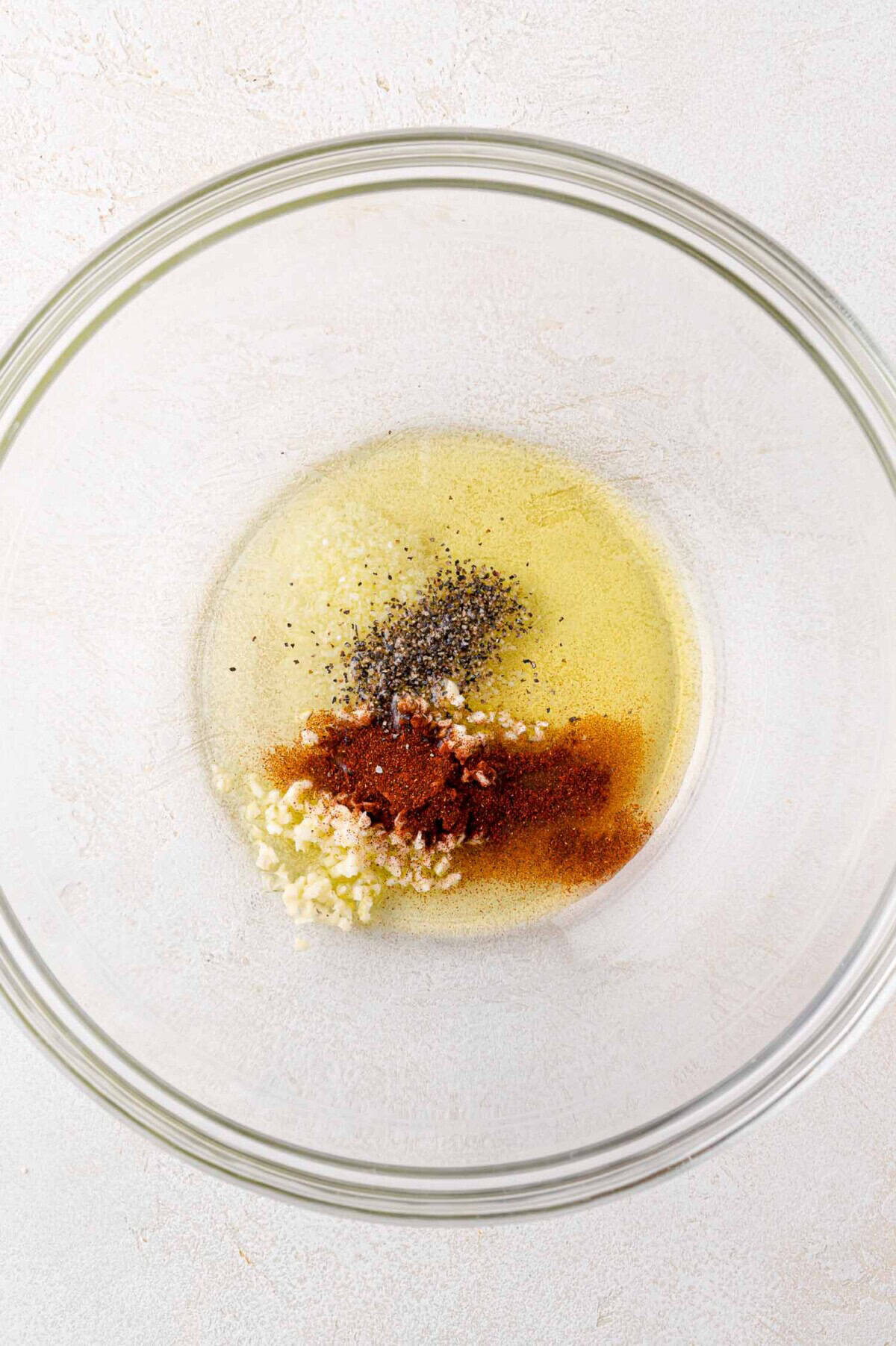 A clear glass bowl containing oil, minced garlic, black pepper, and paprika on a light-colored surface.