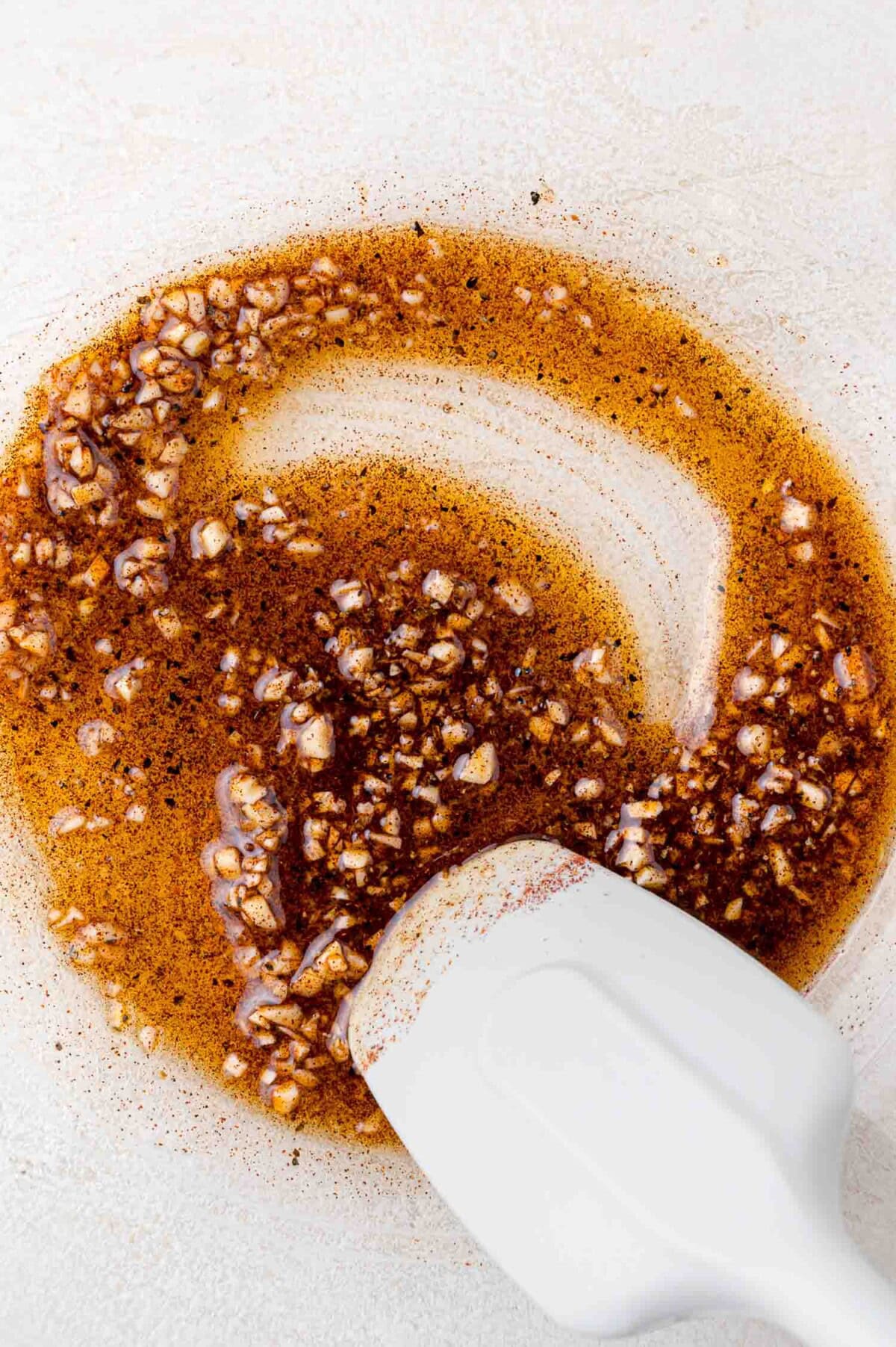 A white spatula mixing a brown liquid with chopped garlic and spices in a clear glass bowl on a light surface.