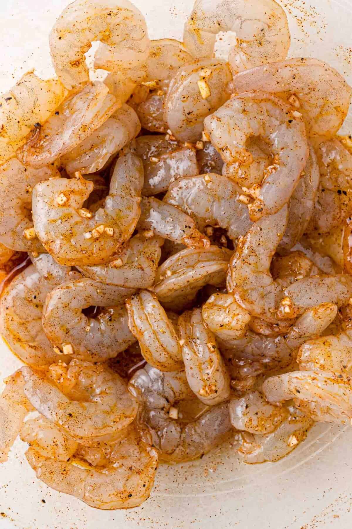 Raw shrimp marinated with spices and minced garlic in a clear bowl, ready for cooking.