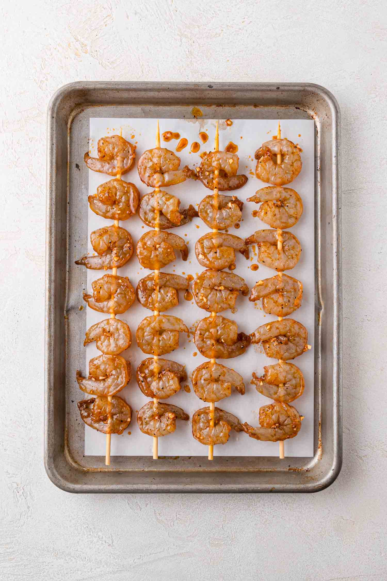 A baking sheet lined with parchment paper holds six skewers, each threaded with five raw, marinated shrimp, arranged in neat rows.