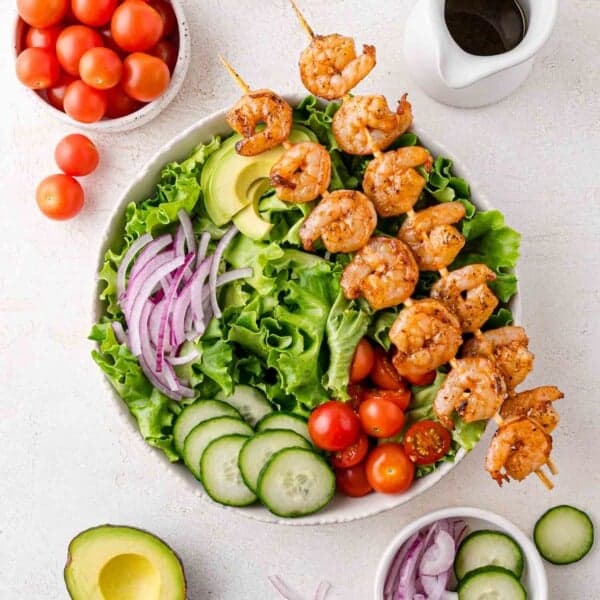 A bowl of salad with grilled shrimp skewers, lettuce, sliced avocado, cucumber, red onion, and cherry tomatoes, with a small jug of dressing and extra vegetables on the side.