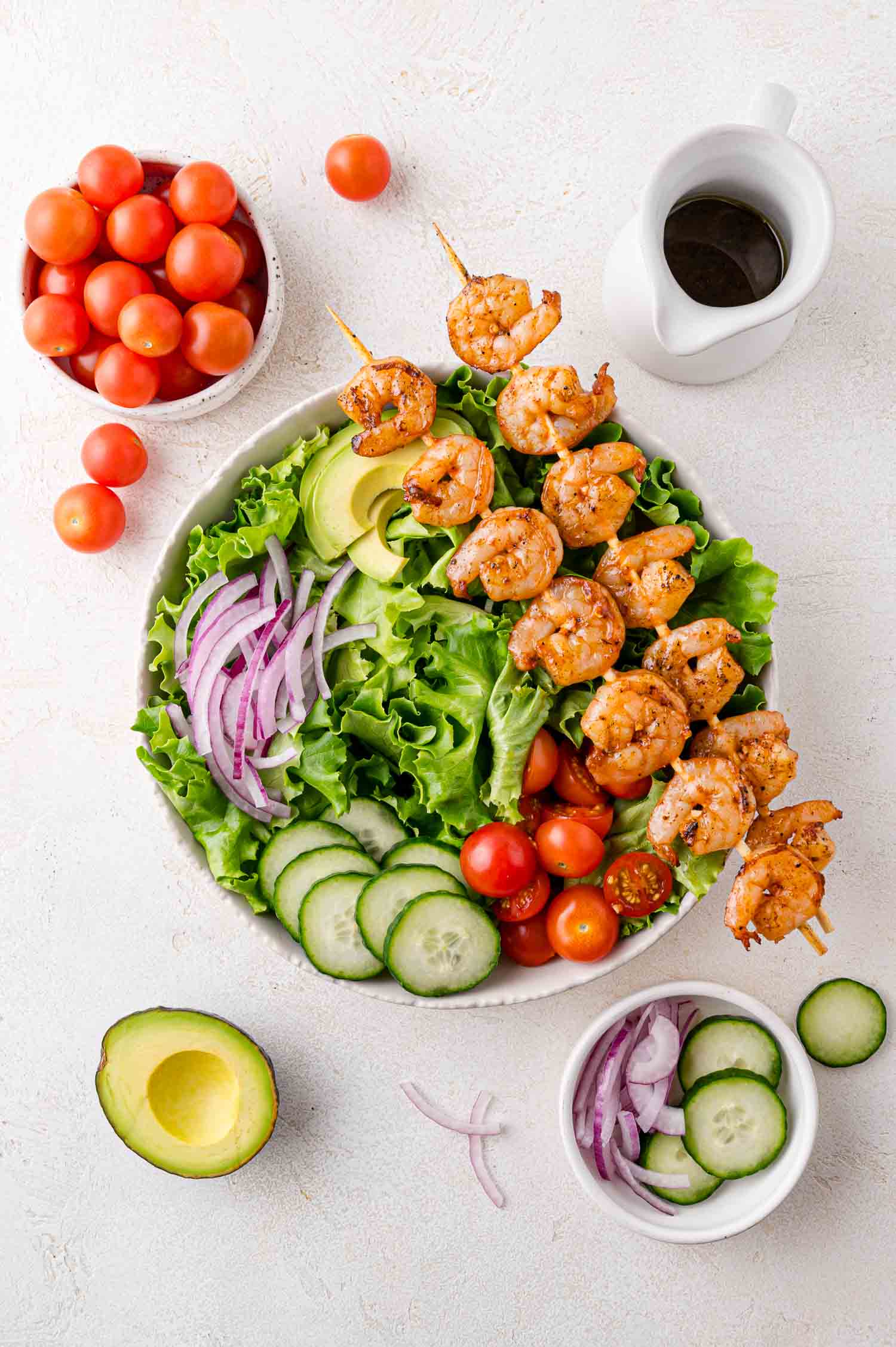 A bowl of salad with skewered grilled shrimp, lettuce, avocado, cucumber, cherry tomatoes, and red onion, surrounded by more tomatoes, avocado, onion, and a jug of dressing.