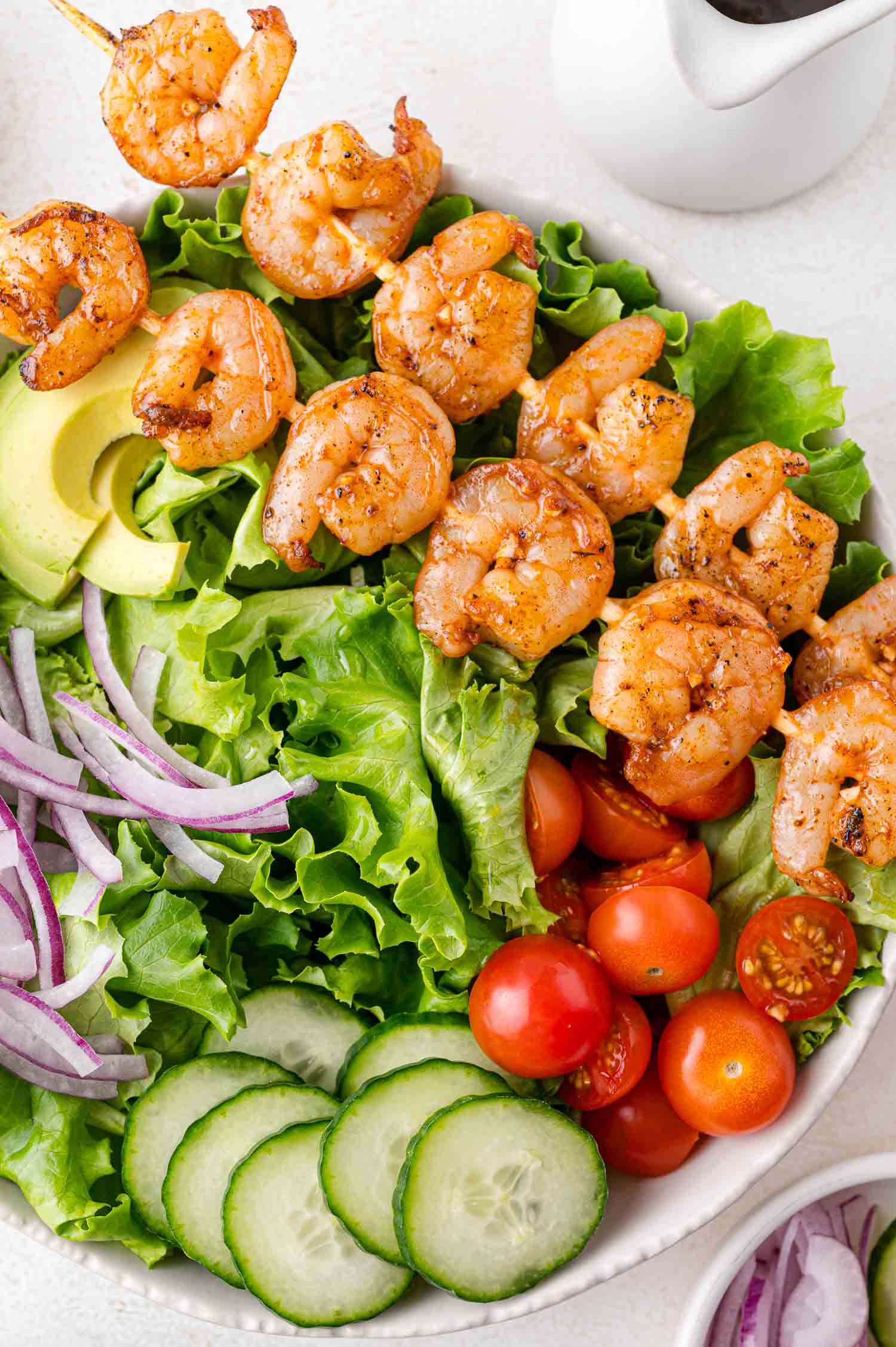 A salad bowl with grilled shrimp skewers, lettuce, avocado slices, red onion, cucumber, and cherry tomatoes.