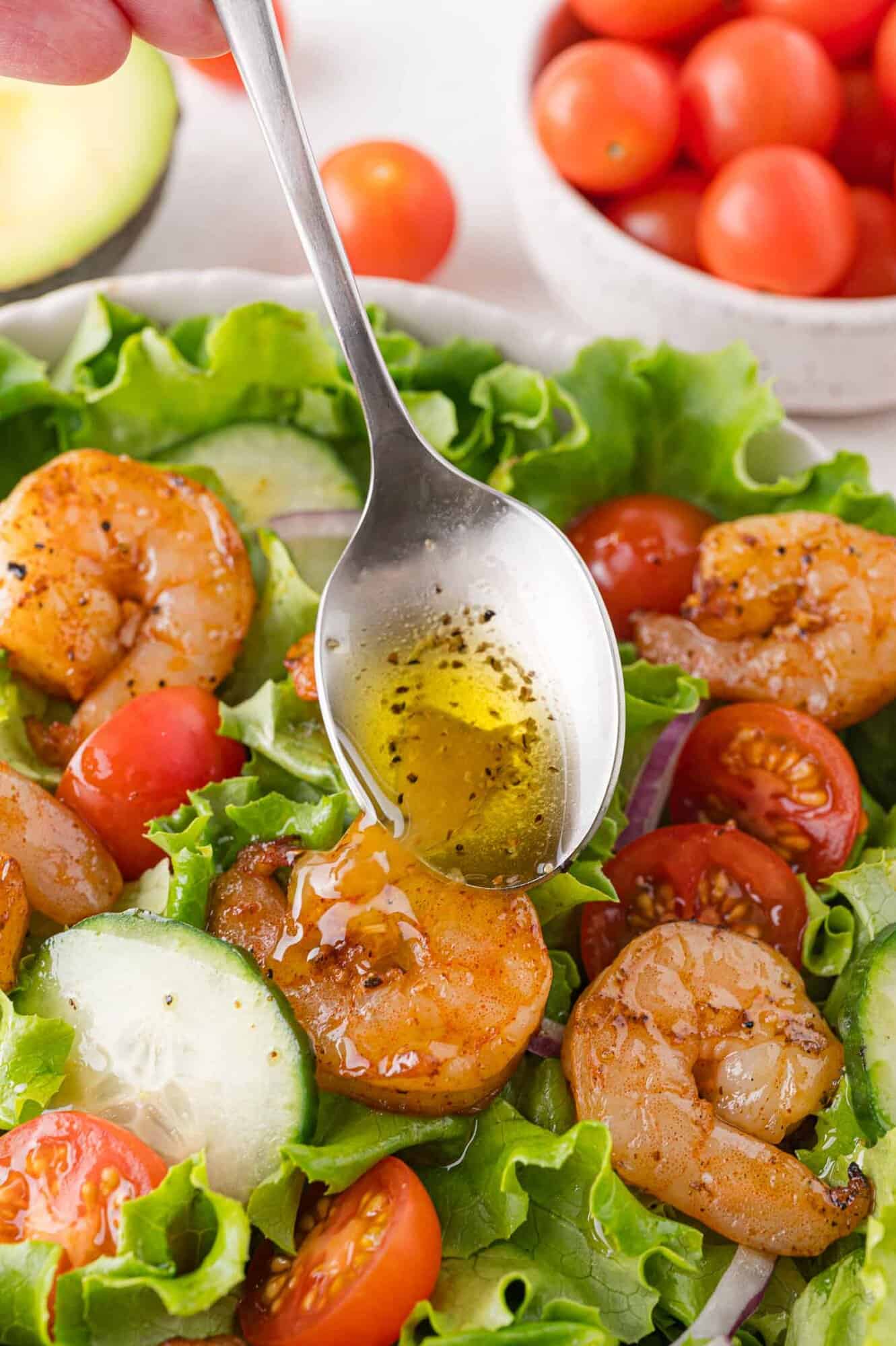 A spoon drizzles vinaigrette over a salad with shrimp, lettuce, cucumber, and cherry tomatoes.