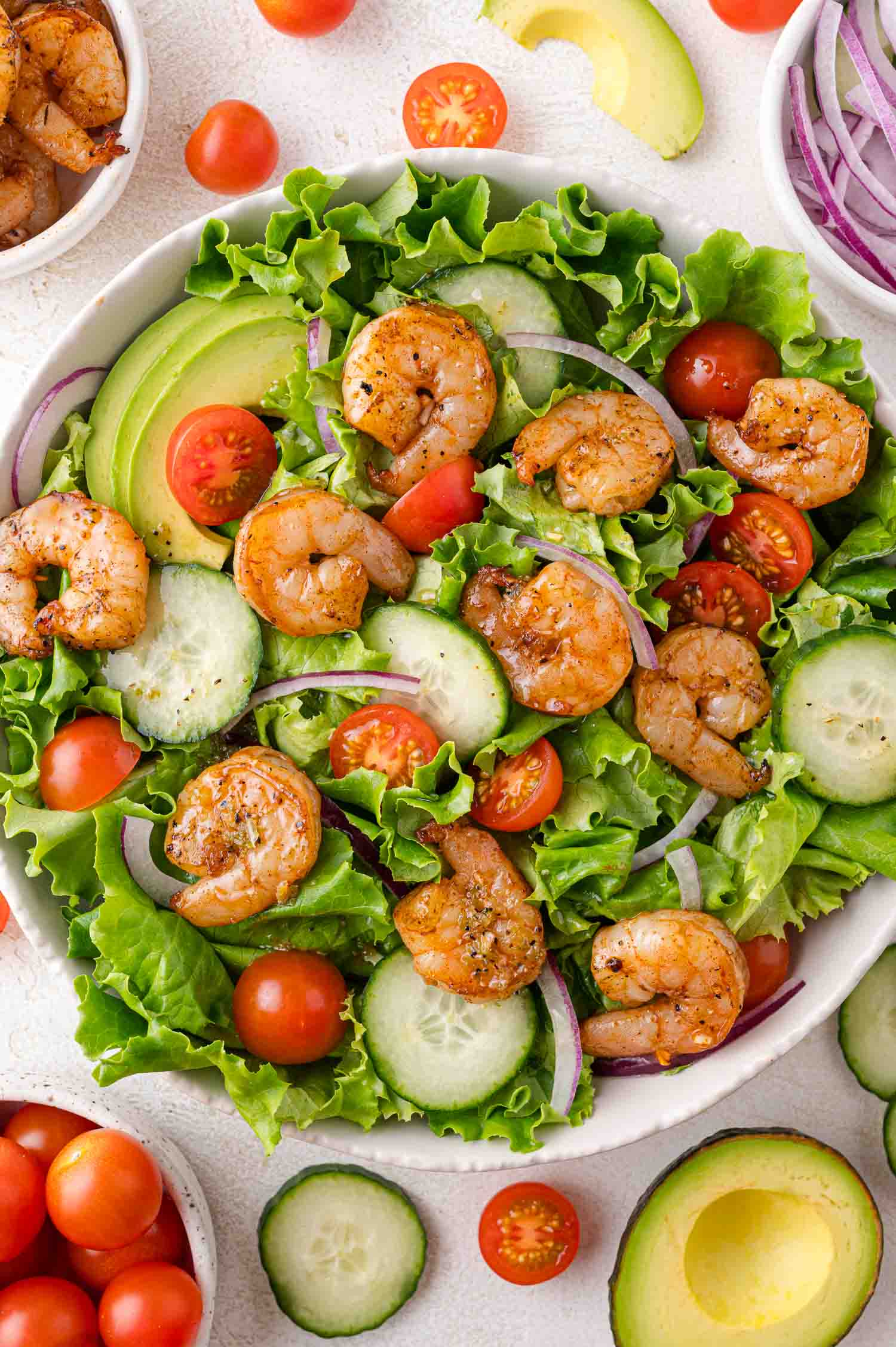 A salad with grilled shrimp, lettuce, cucumber slices, cherry tomatoes, avocado, and red onion in a white bowl, surrounded by salad ingredients.