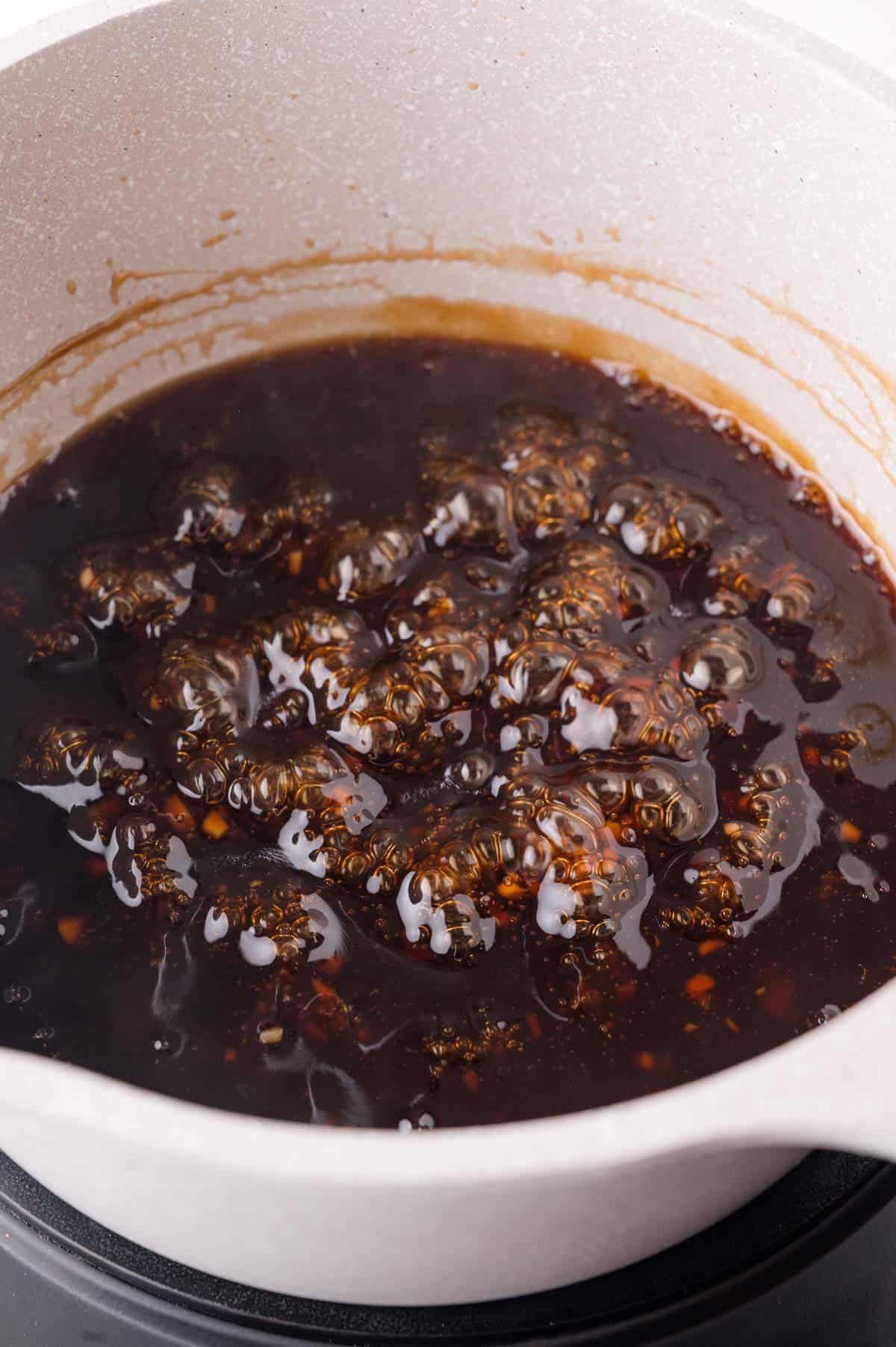 A close-up of a pot on a stove with a dark brown sauce bubbling and simmering.
