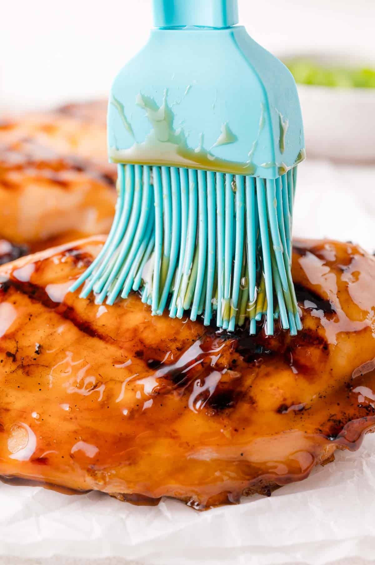 A silicone brush applies glaze to a grilled chicken breast on parchment paper.