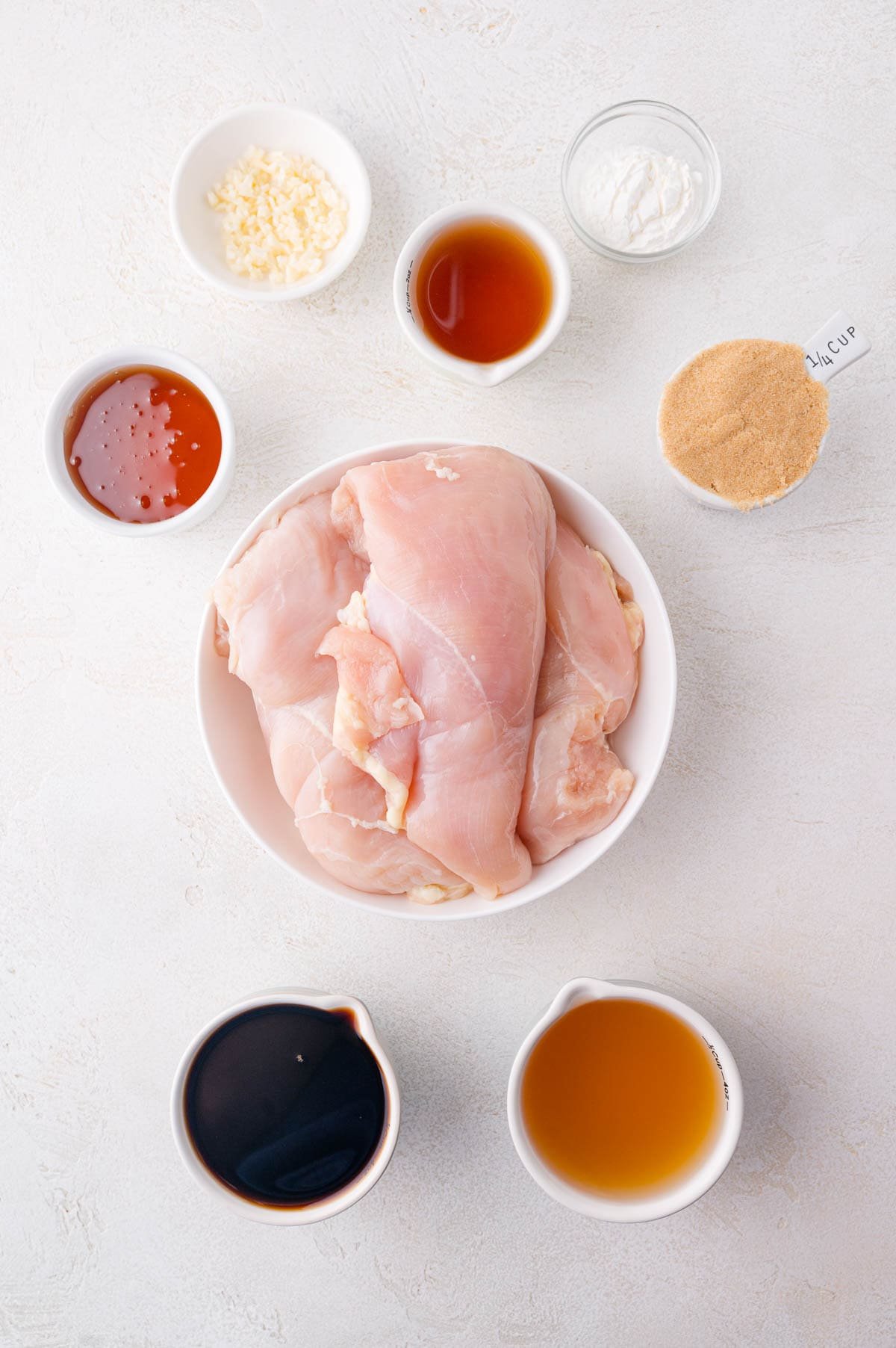 A bowl of raw chicken breasts surrounded by small bowls containing minced garlic, honey, soy sauce, brown sugar, apple cider vinegar, cornstarch, and another brown liquid on a white surface.