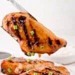 A grilled chicken breast glazed with sauce, topped with sesame seeds and chopped green onions, held by tongs above a plate of rice.