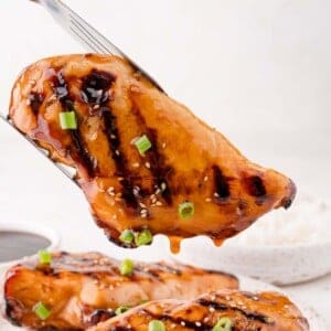 A grilled chicken breast glazed with sauce, topped with sesame seeds and chopped green onions, held by tongs above a plate of rice.