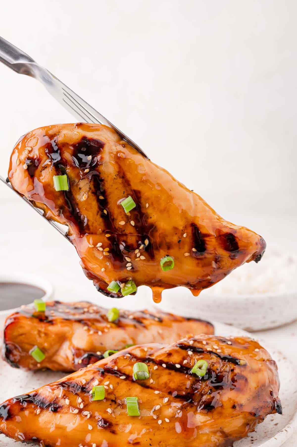 A grilled chicken breast glazed with sauce and garnished with sesame seeds and chopped green onions held by tongs above two similar pieces on a plate.