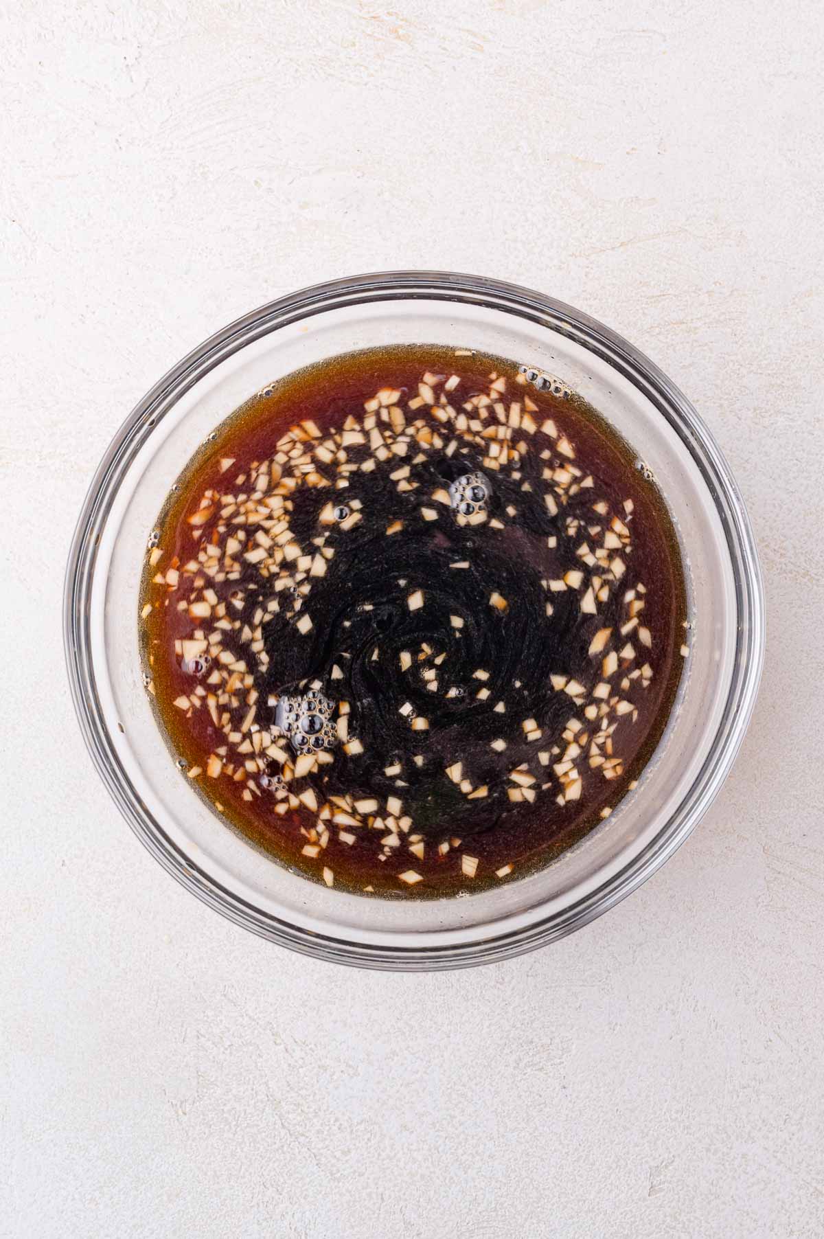 A clear glass bowl containing a dark liquid with finely chopped garlic pieces floating on the surface, set on a light-colored background.