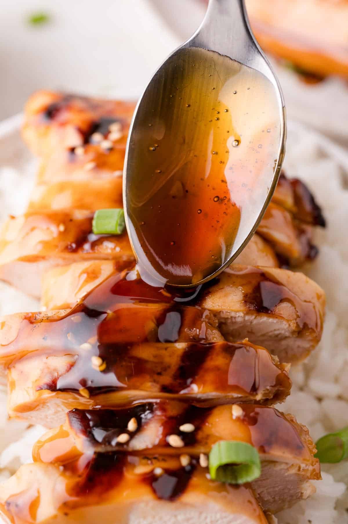 A spoon drizzles teriyaki sauce over sliced grilled chicken breast on a bed of white rice, garnished with sesame seeds and chopped green onions.