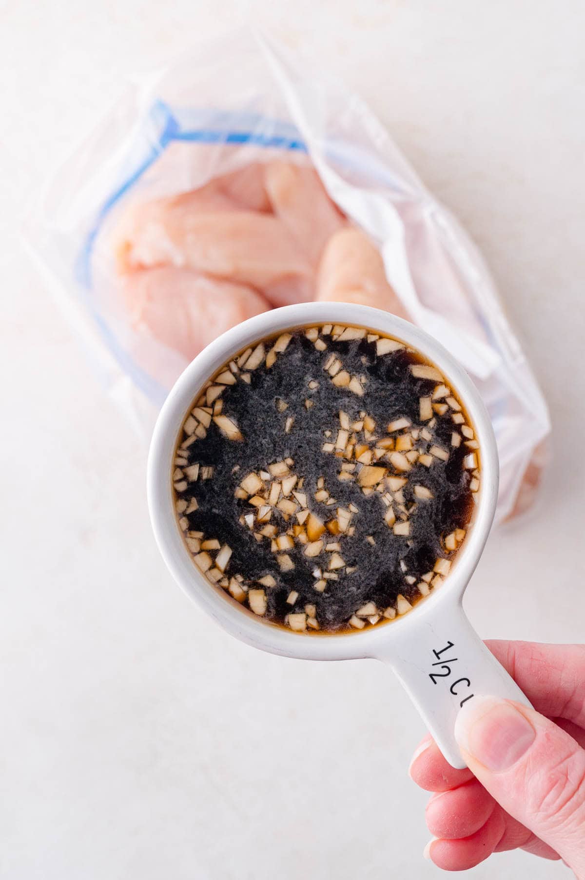 A hand holds a half-cup measuring cup filled with a chopped garlic and soy sauce mixture above a plastic bag containing raw chicken breasts.