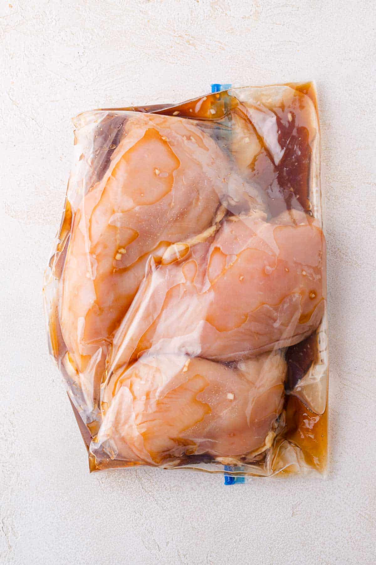 Raw chicken breasts marinating in a sealed plastic bag with a brown liquid marinade, placed on a light textured surface.