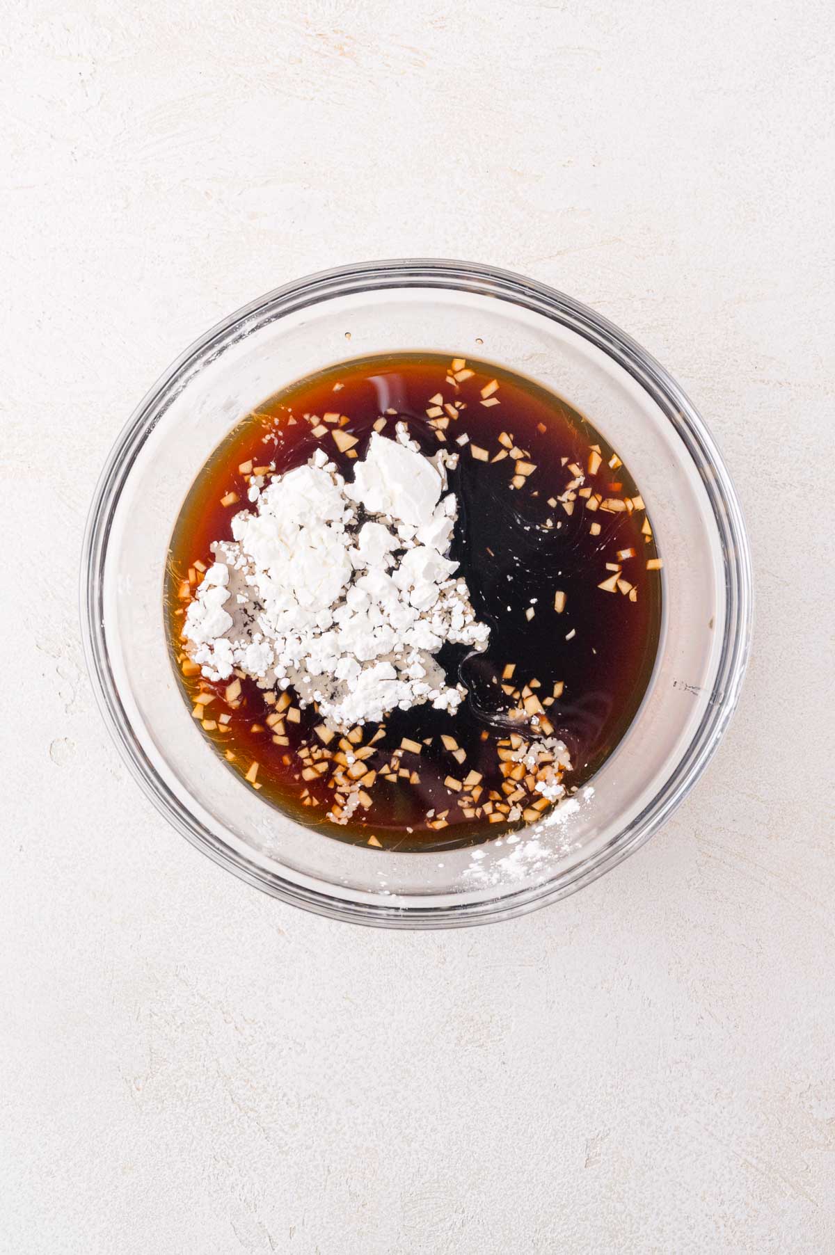 A glass bowl containing a mixture of soy sauce, minced garlic, and cornstarch on a light-colored surface.