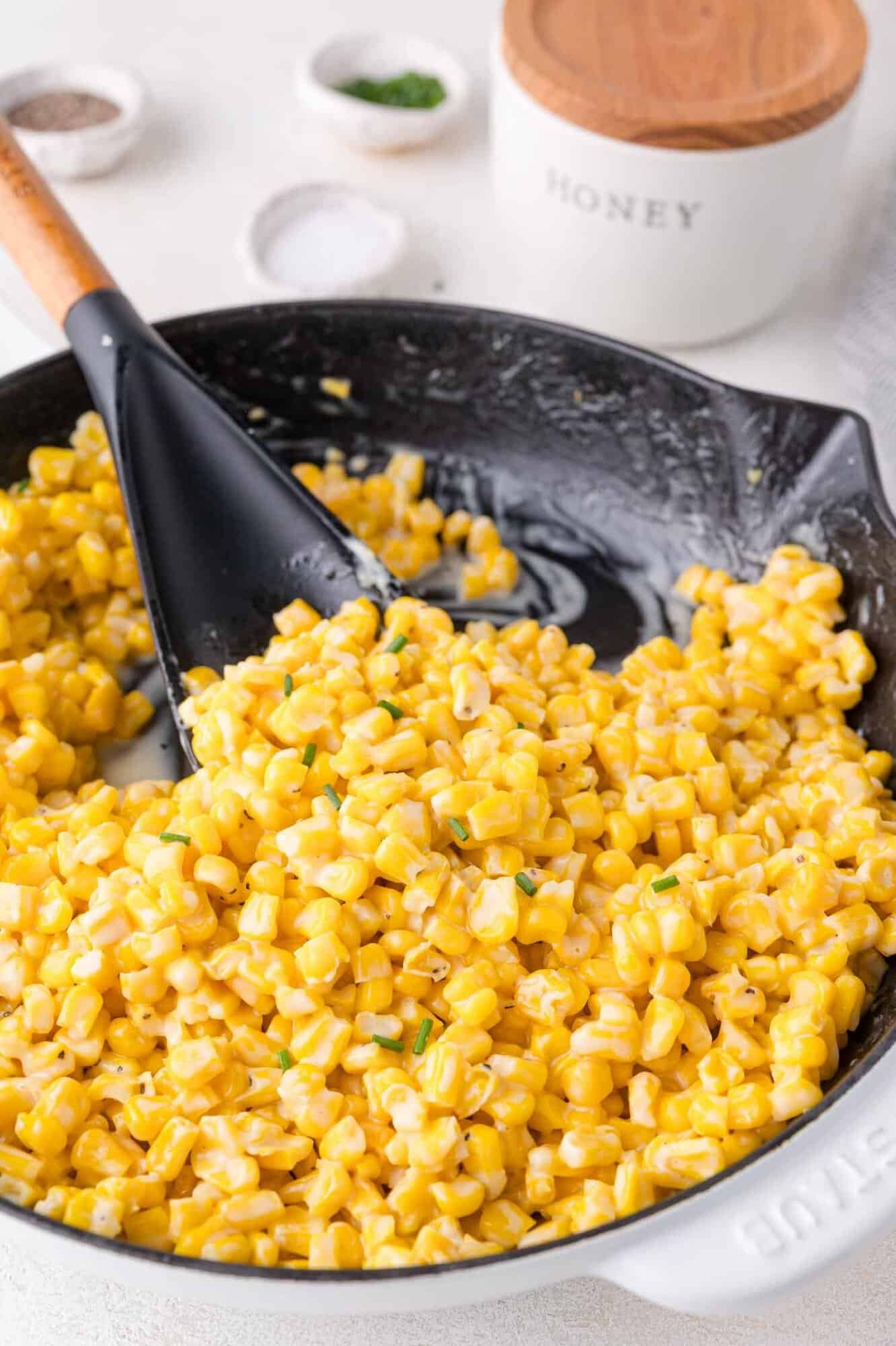 A skillet filled with creamy honey butter skillet corn, mixed with herbs, and a black spatula resting inside. Salt, pepper, and a container labeled Honey are in the background.
