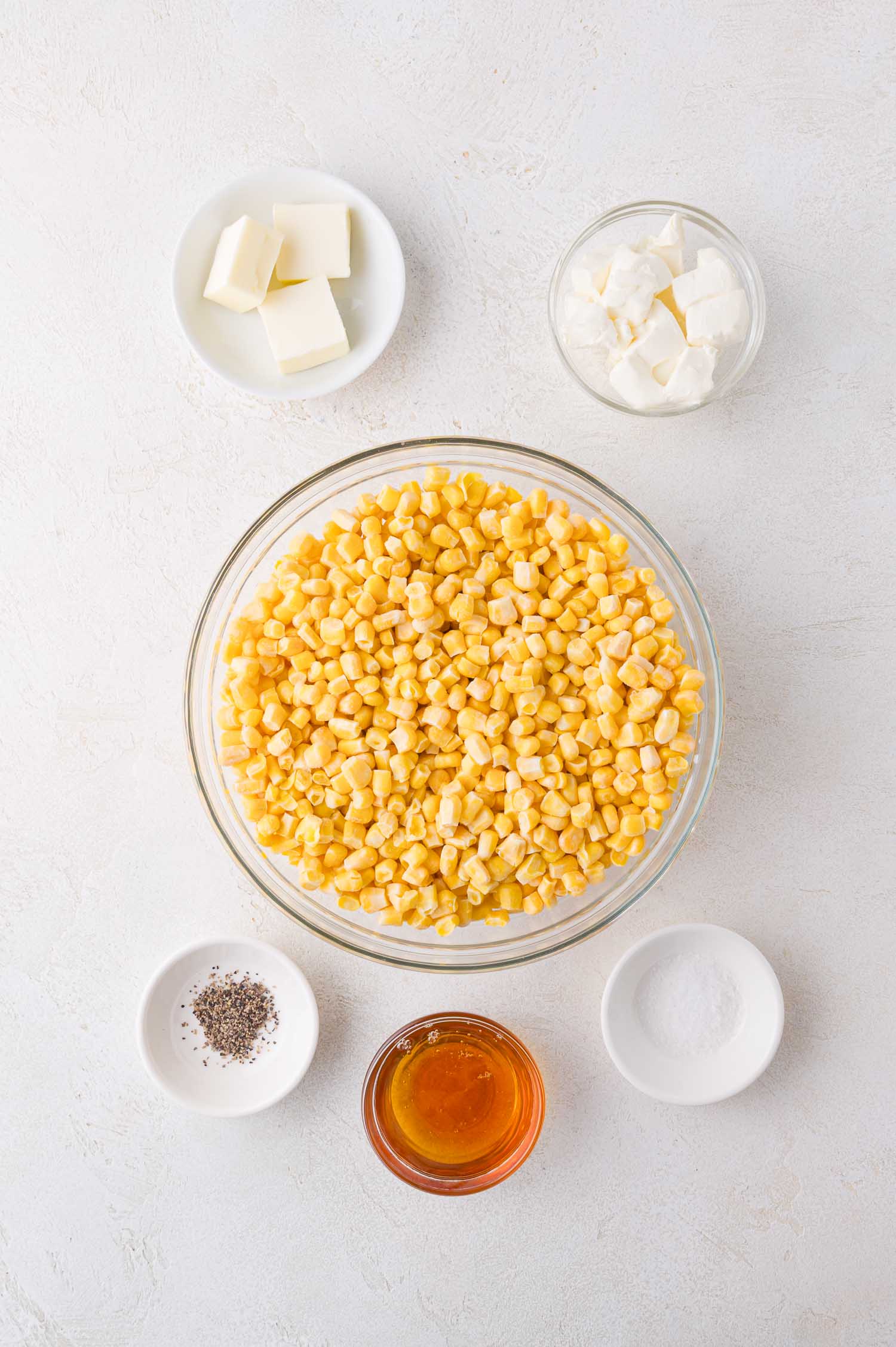 A bowl of corn kernels surrounded by small bowls of butter, sour cream, salt, pepper, and honey on a white surface.