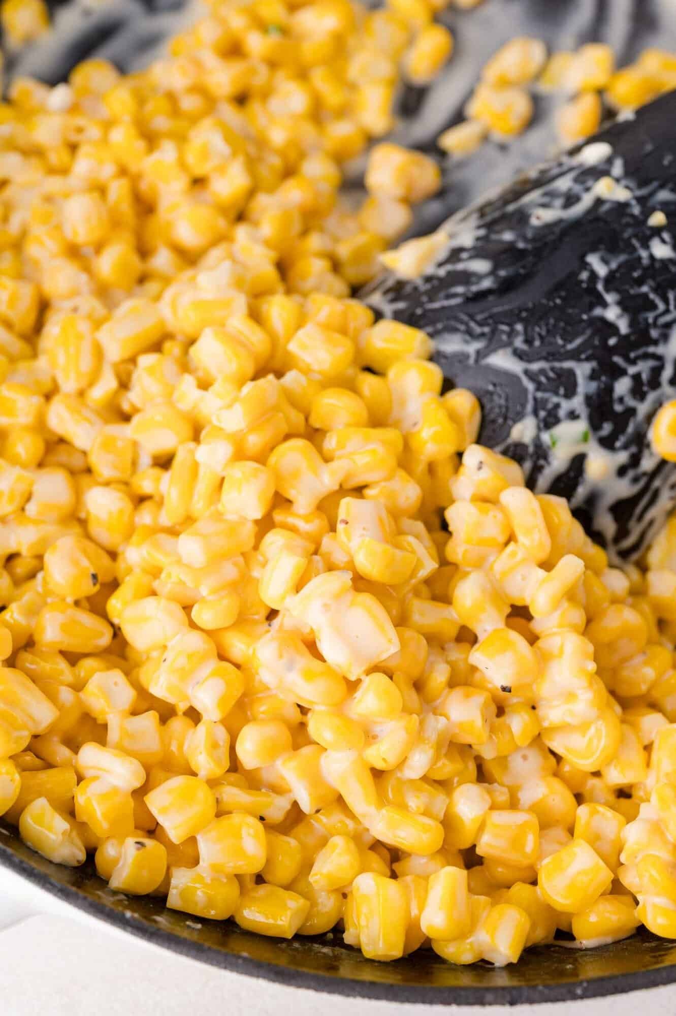Close-up of honey butter skillet corn being stirred in a pan with a black spatula. The corn kernels are coated in a creamy sauce.
