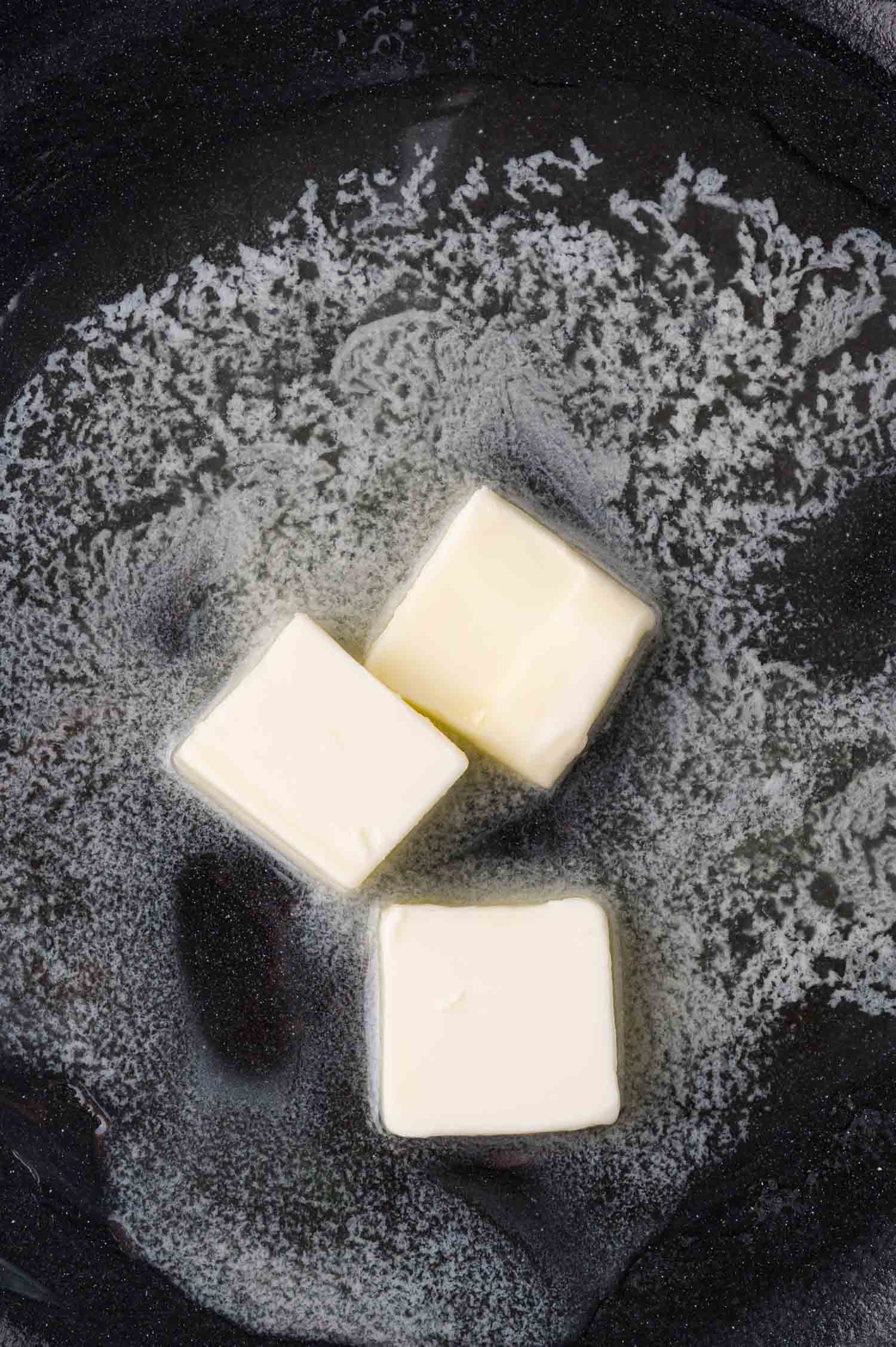 Three cubes of butter are melting in a black pan, creating a pool of liquid butter around them.