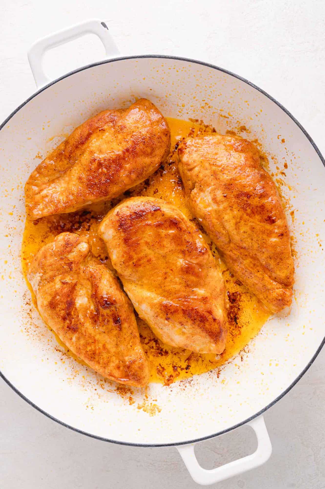 Four cooked, seasoned chicken breasts in a white skillet with visible juices and some browning.