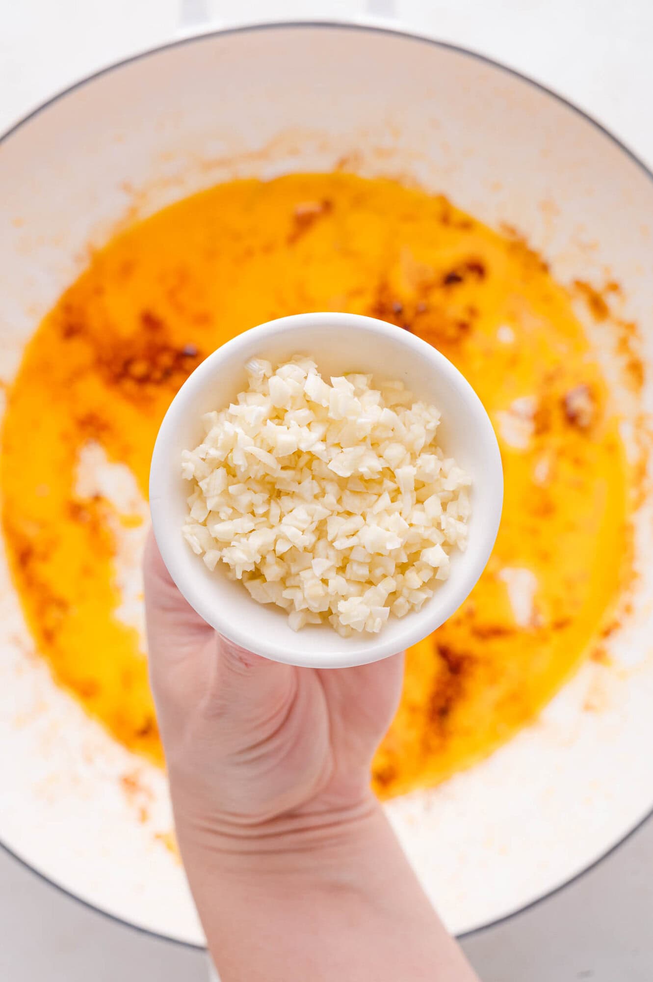 A hand holds a bowl of finely chopped garlic above a pan containing oil and spices.
