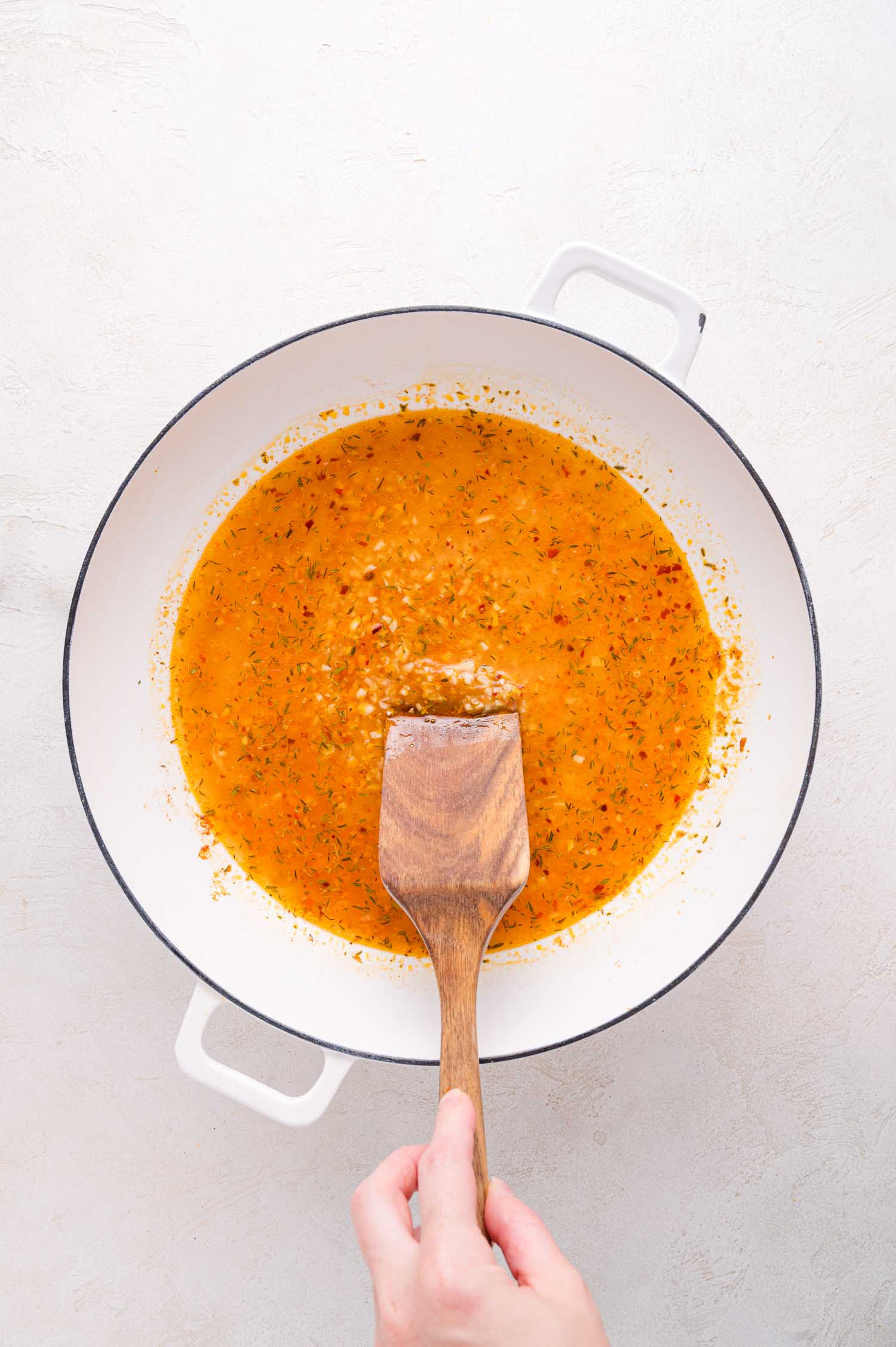 A hand stirs an orange-colored sauce with herbs in a white pot using a wooden spatula.