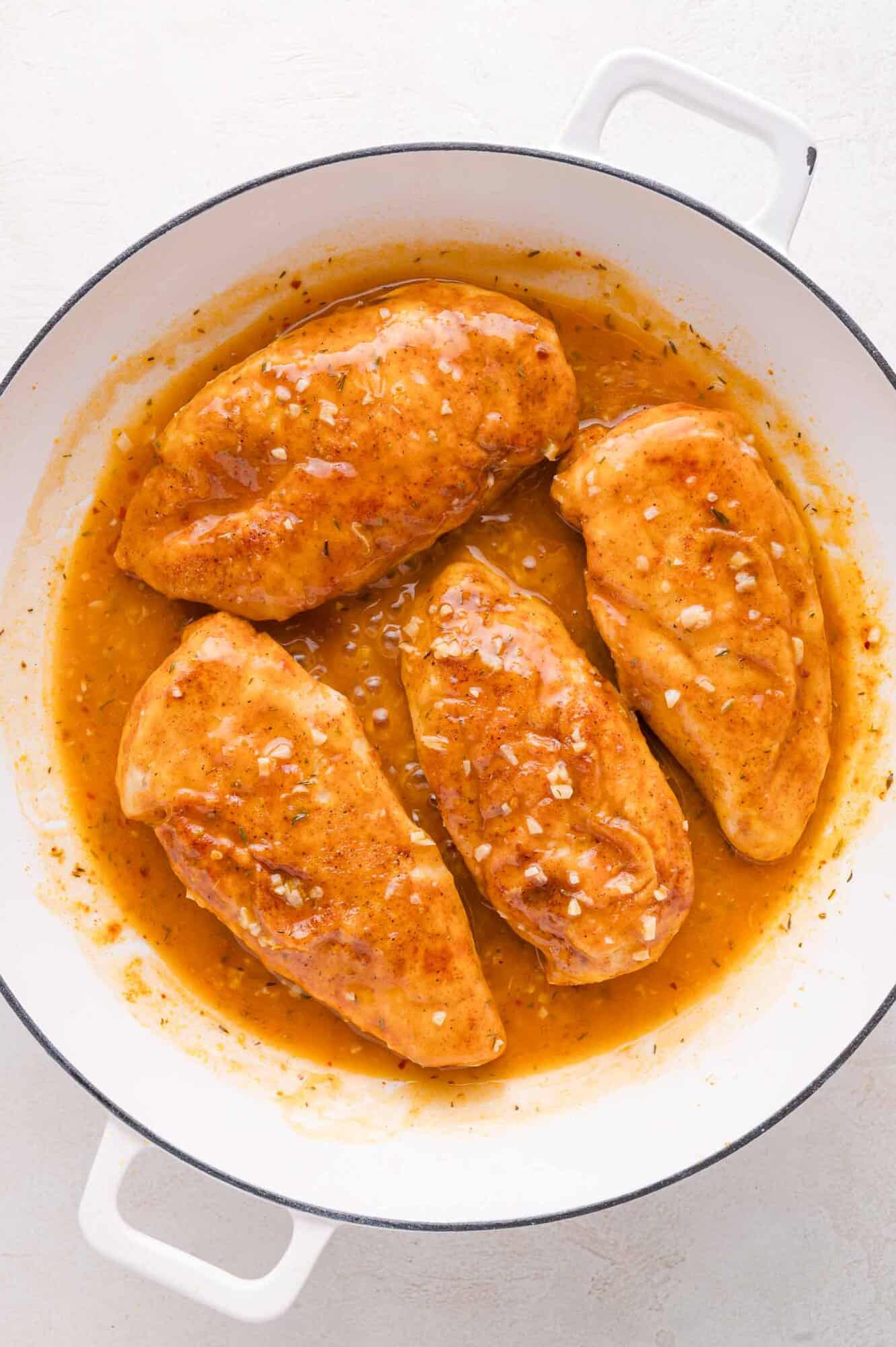 Four seasoned chicken breasts simmer in a light brown sauce inside a white skillet, with visible bits of diced garlic in the sauce.