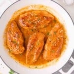 Four cooked chicken breasts in a skillet, covered in a thick, orange-brown garlic butter sauce and garnished with chopped herbs.