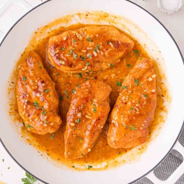Four cooked chicken breasts in a skillet, covered in a thick, orange-brown garlic butter sauce and garnished with chopped herbs.