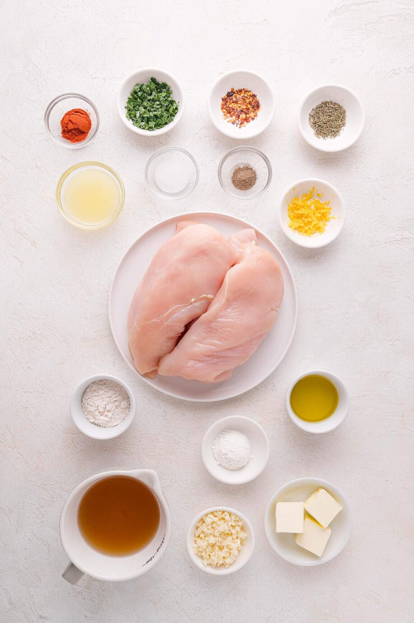 A plate with two raw chicken breasts surrounded by small bowls containing spices, herbs, lemon zest, flour, broth, garlic, butter, and oil on a white surface.