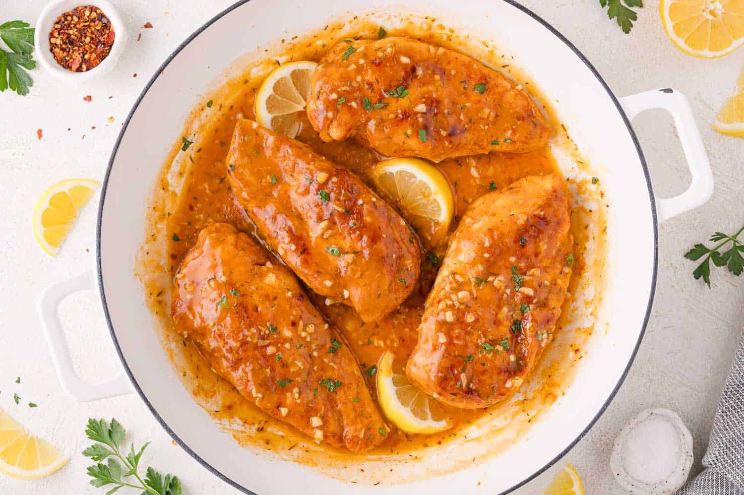 Four cooked chicken breasts in a white pan with a golden lemon-garlic sauce, garnished with lemon slices and chopped herbs.