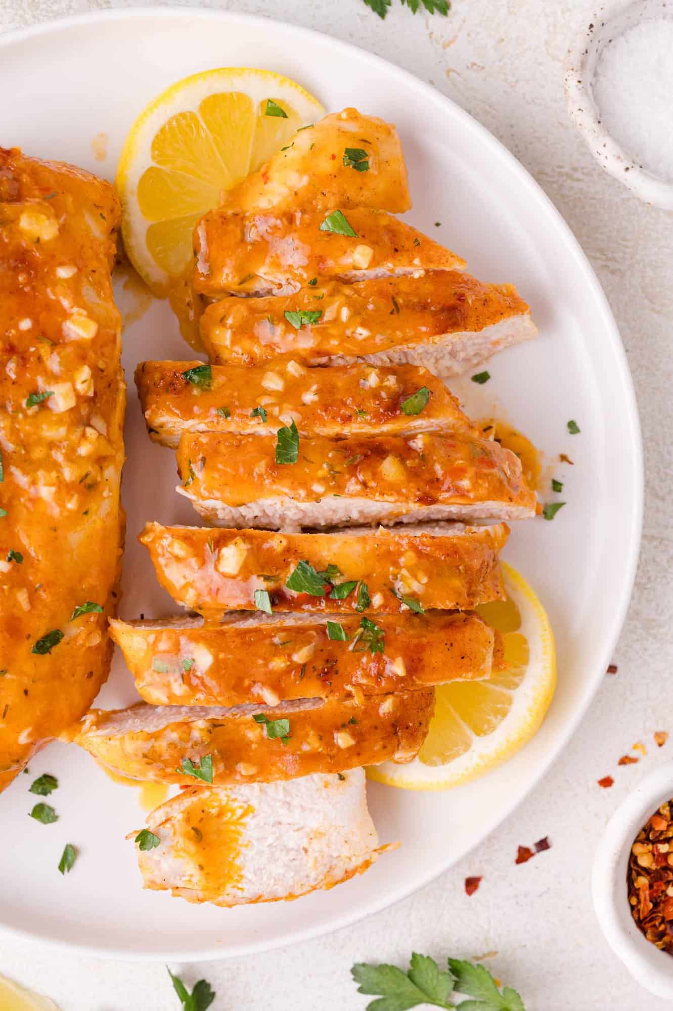 Sliced cooked chicken breast topped with a garlic and herb sauce, garnished with chopped parsley and lemon slices on a white plate.