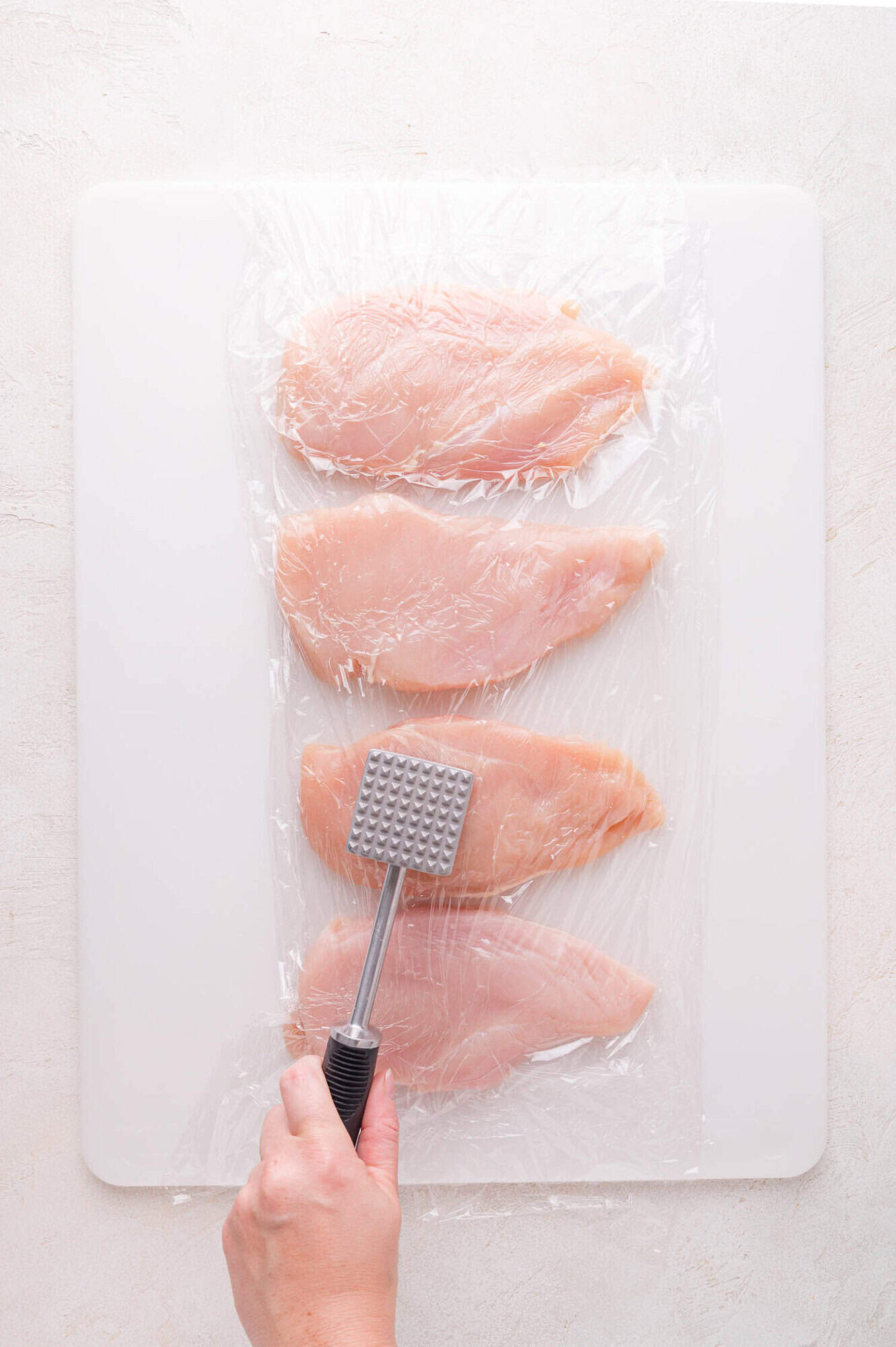Three raw chicken breasts on a cutting board covered with plastic wrap, with a hand holding a meat mallet above them, ready to tenderize.