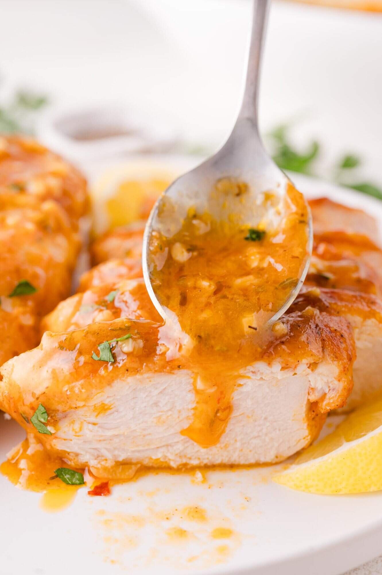A spoon pours orange sauce over a sliced piece of cooked chicken breast, garnished with herbs, on a white plate with a lemon wedge.