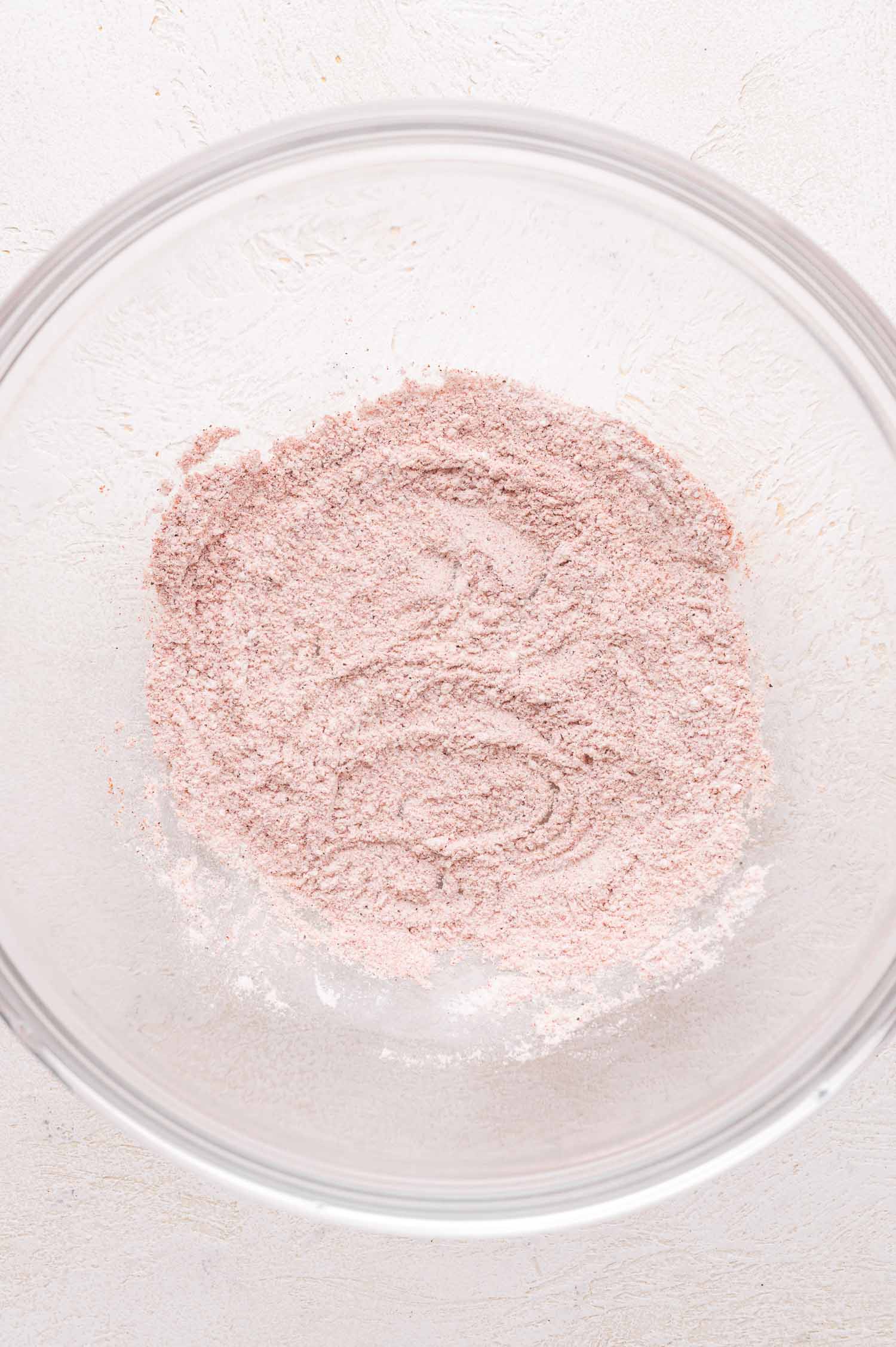 A clear glass bowl containing a light pinkish-brown spice mixture on a white textured surface.