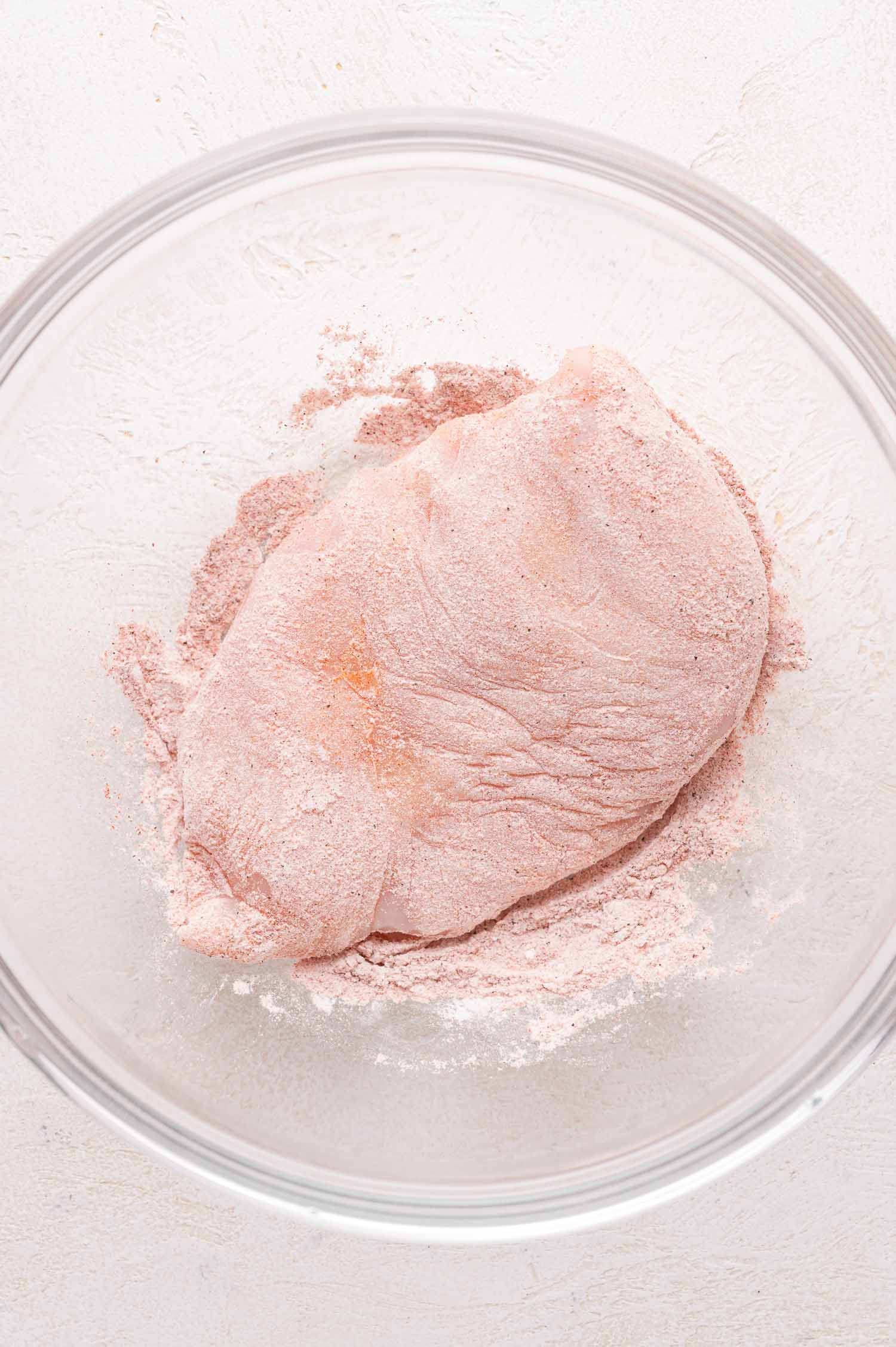 Raw chicken breast coated with a pink seasoning blend in a glass bowl, placed on a light-colored surface.