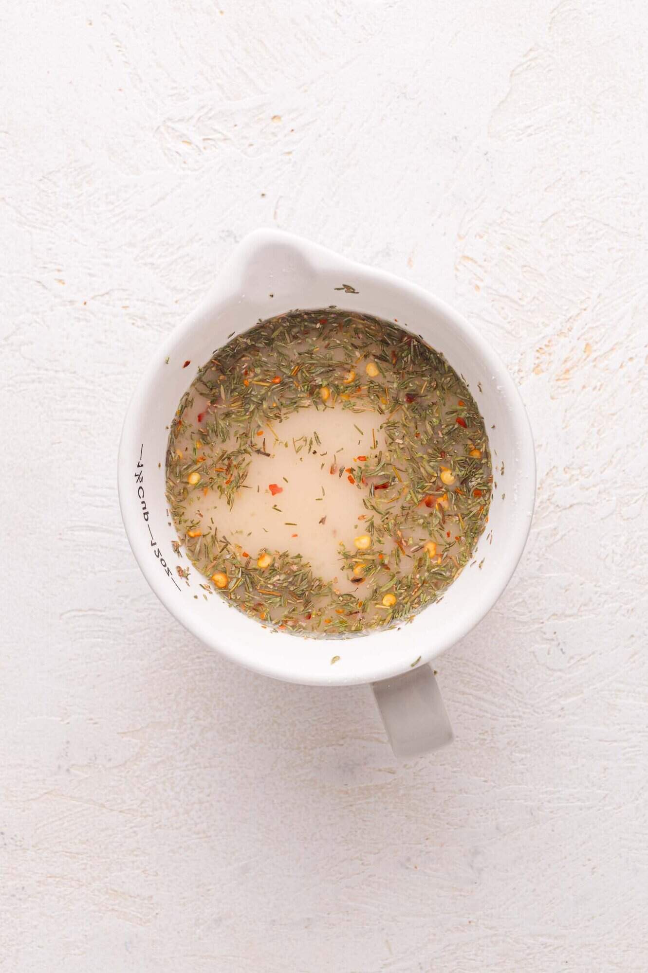 A white measuring cup filled with a sauce containing herbs and spices, viewed from above on a light textured surface.