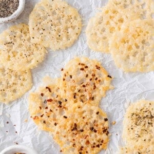 Several round parmesan crisps with spices, such as black pepper and red pepper flakes, are arranged on crinkled white parchment paper.