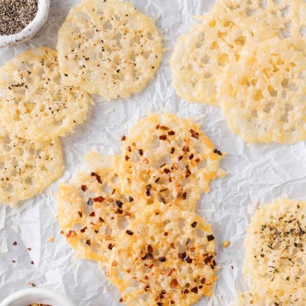 Several round parmesan crisps with spices, such as black pepper and red pepper flakes, are arranged on crinkled white parchment paper.
