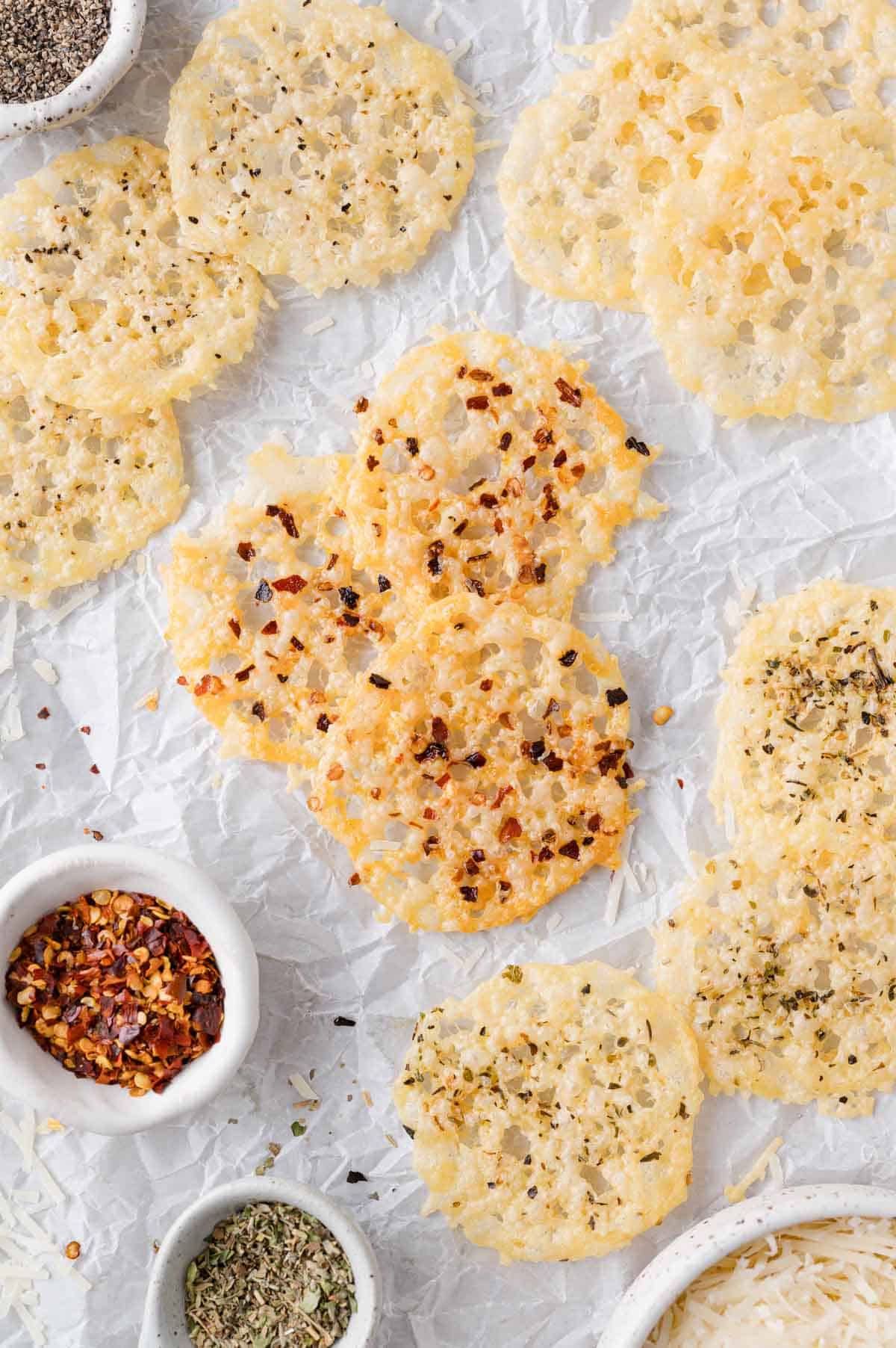 Crispy cheese rounds rest on crumpled parchment paper, surrounded by small bowls containing chili flakes, dried herbs, and shredded cheese.