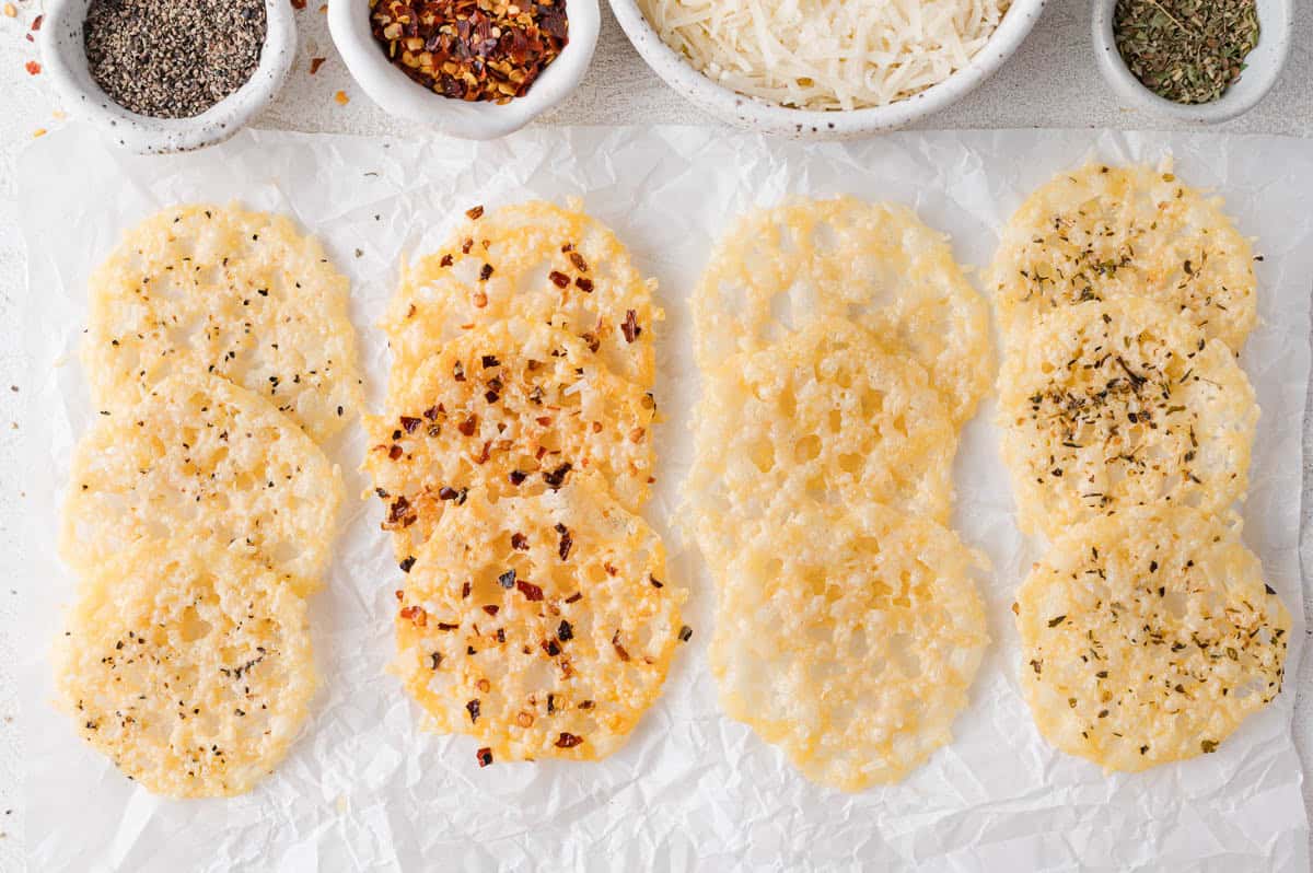 Four rows of cheese crisps on parchment paper, each topped with different seasonings; bowls of shredded cheese, black pepper, red pepper flakes, and herbs are above.