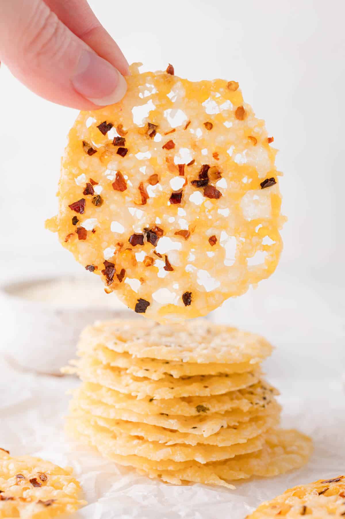 A hand holds up a thin, round cheese crisp topped with red pepper flakes above a stack of similar crisps on a white surface.