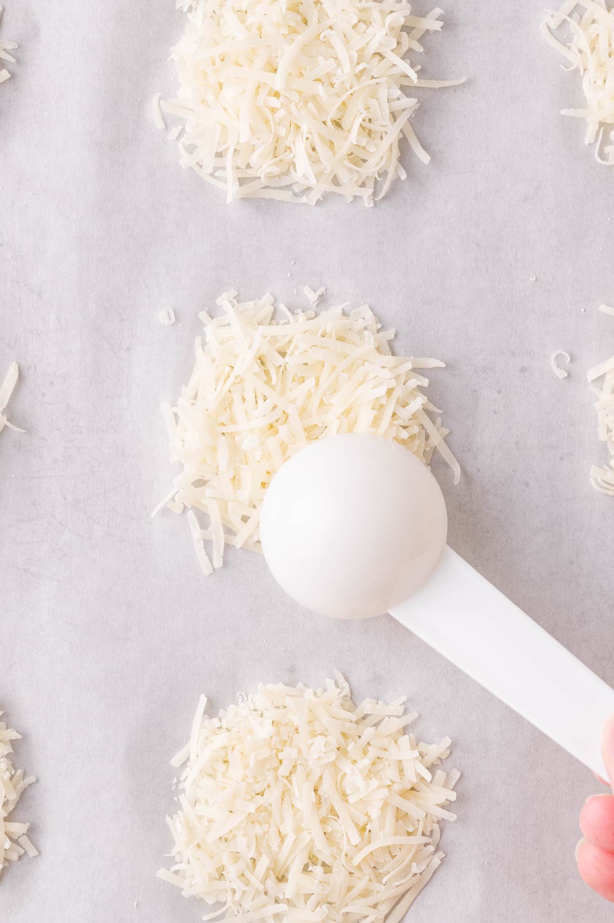 A white measuring spoon holds shredded cheese, forming small piles on parchment paper.