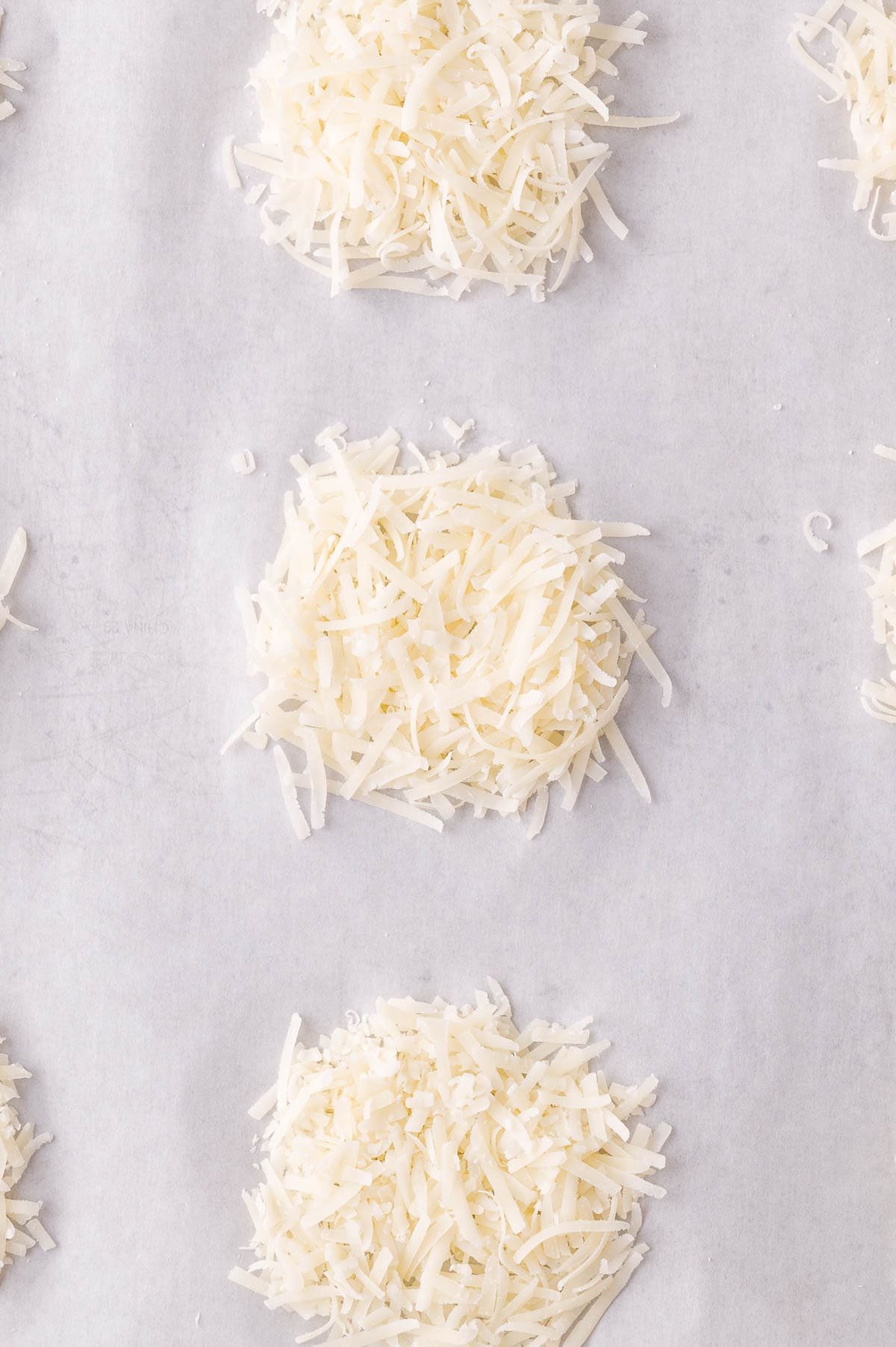 Shredded Parmesan cheese is arranged in small mounds on a sheet of parchment paper, spaced evenly apart.