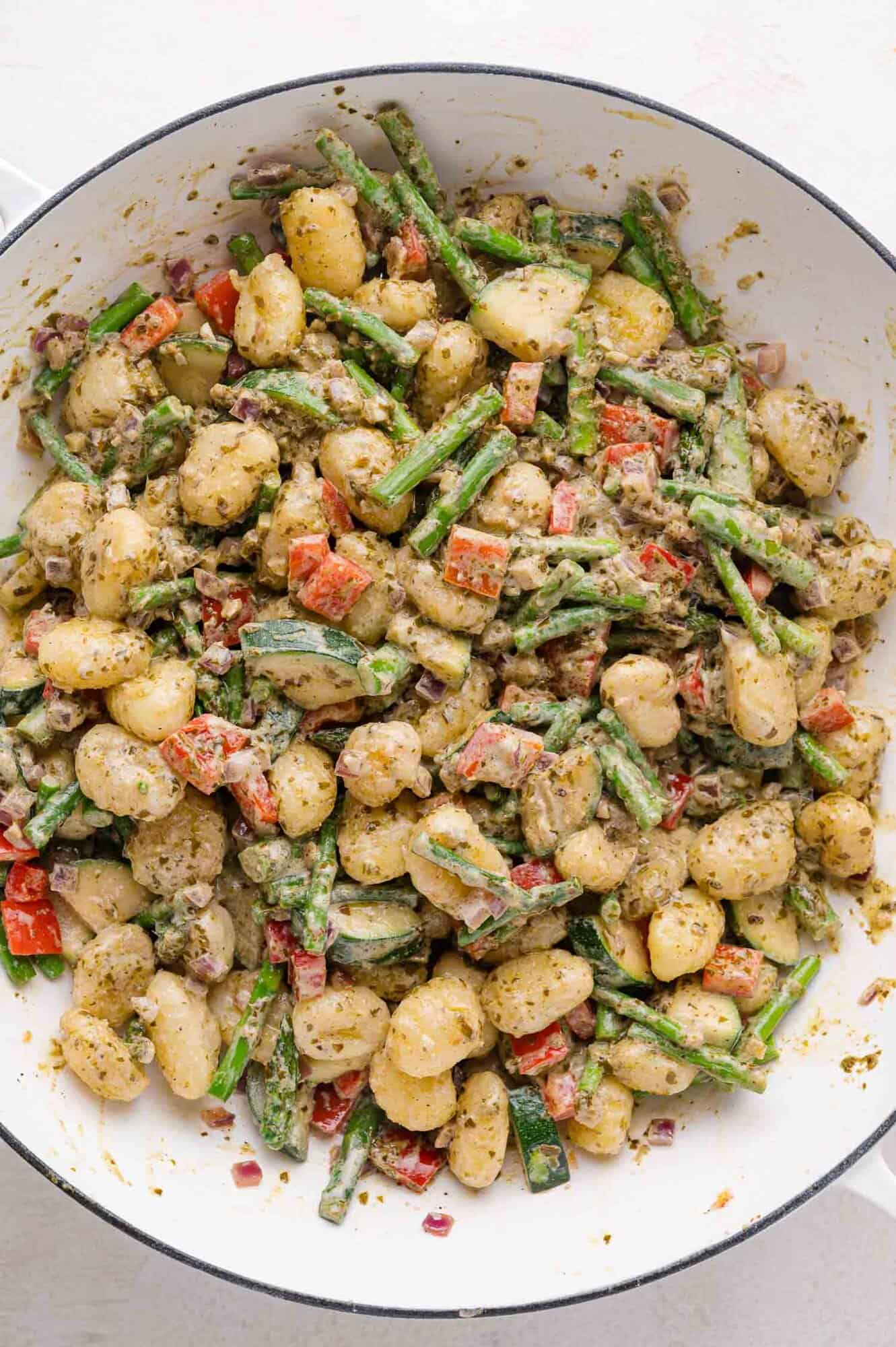 A skillet filled with gnocchi, asparagus, red bell peppers, zucchini, and onions, all coated in a green pesto sauce.