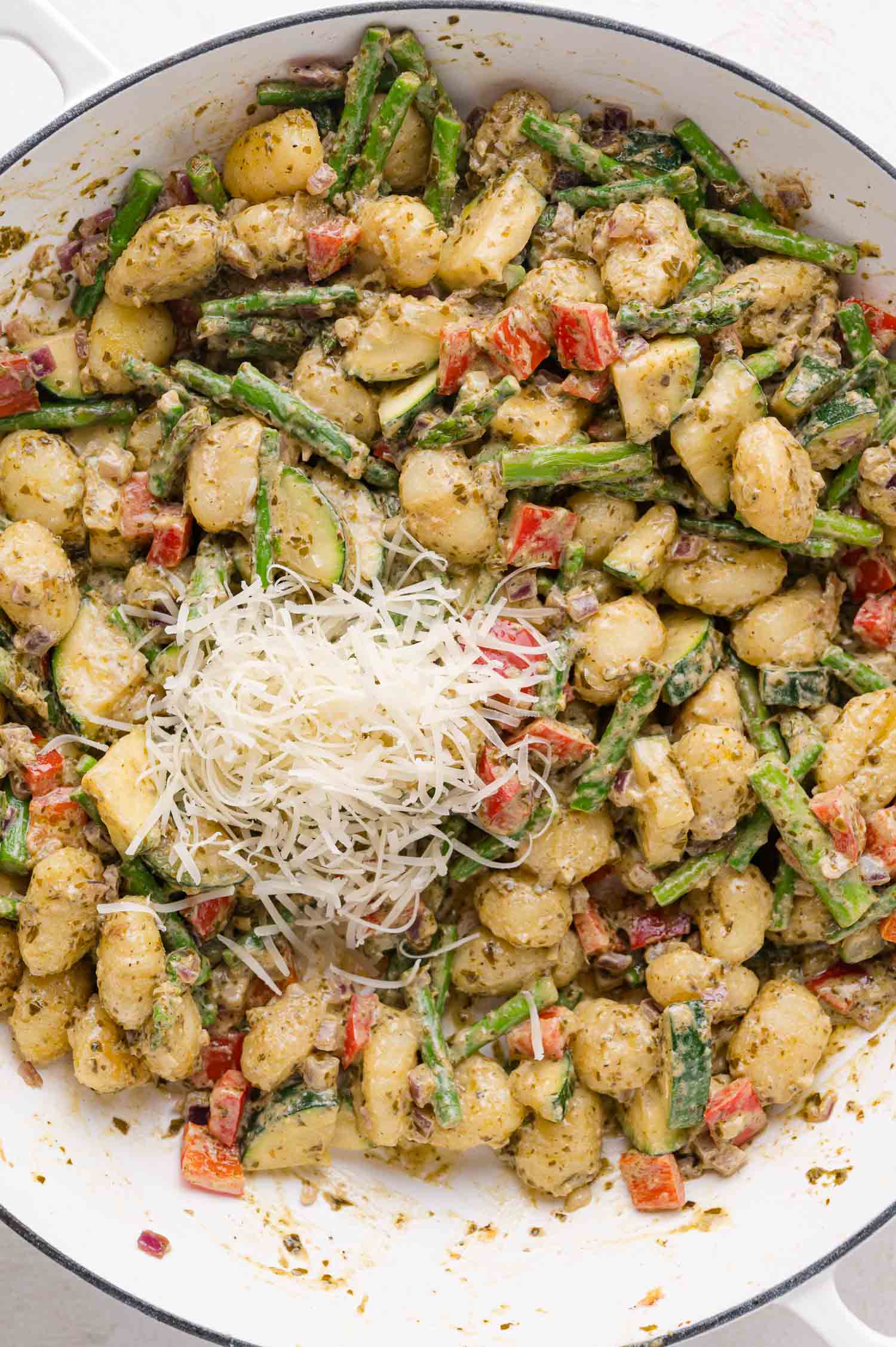 A skillet filled with gnocchi, asparagus, red bell peppers, pesto sauce, and a pile of shredded cheese in the center.