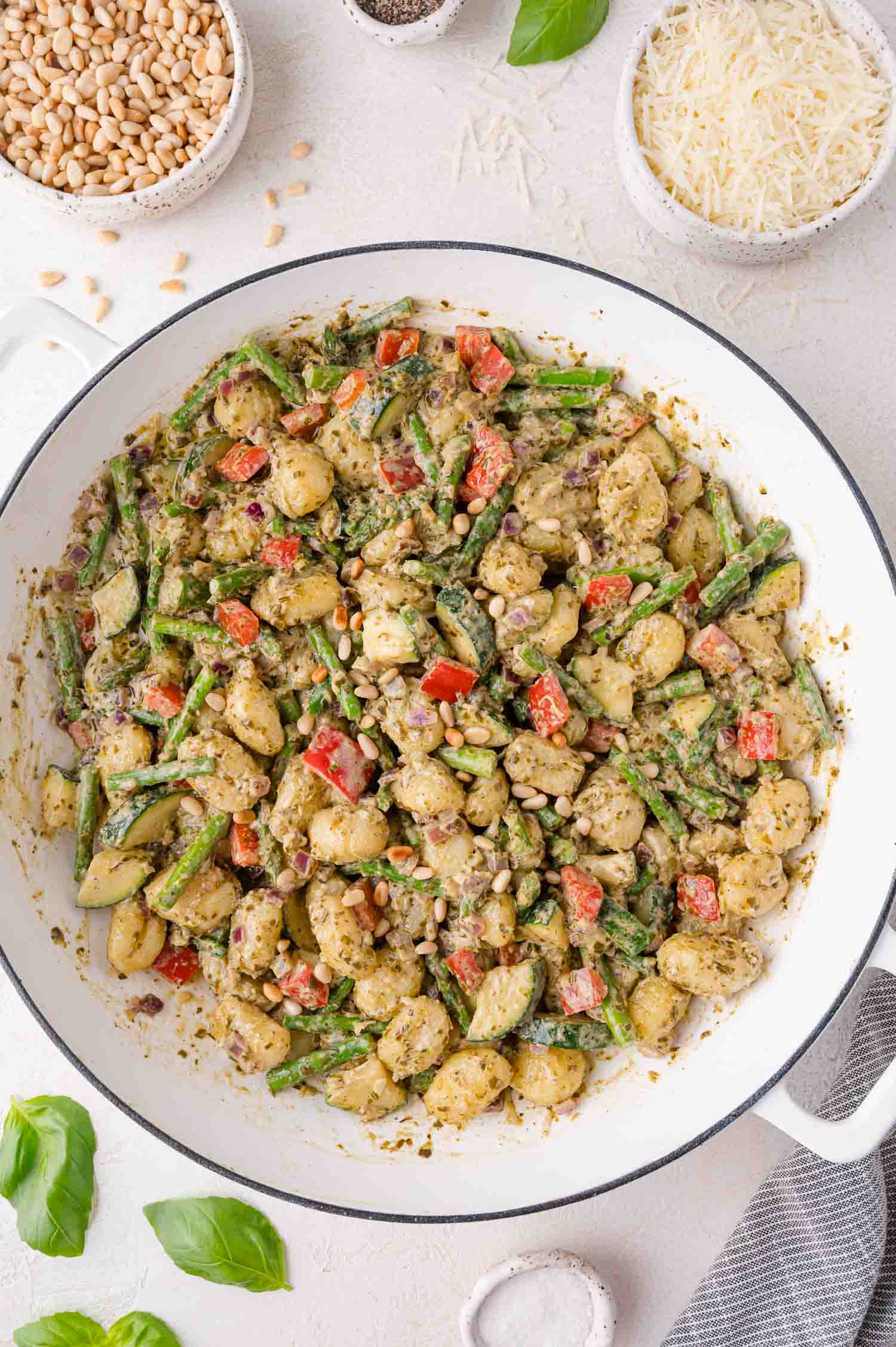 A white skillet filled with gnocchi, asparagus, red bell peppers, and pesto sauce, garnished with pine nuts, on a light surface with bowls of pine nuts and grated cheese nearby.