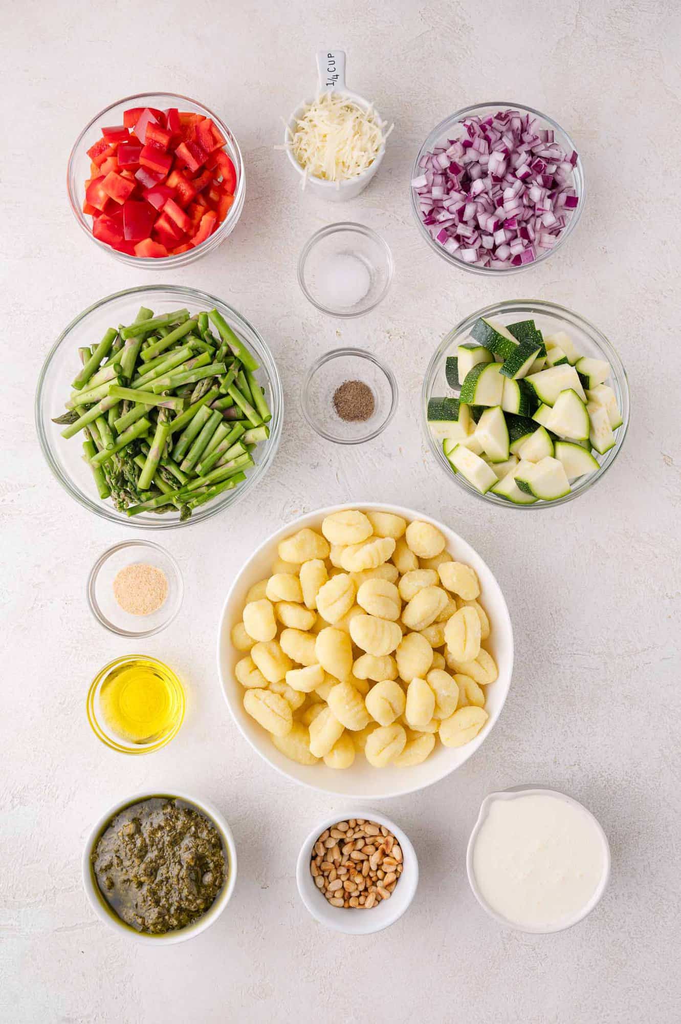 Overhead view of bowls containing gnocchi, chopped red bell pepper, asparagus, red onion, zucchini, pesto, cream, olive oil, pine nuts, shredded cheese, garlic powder, salt, and pepper.