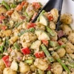 Gnocchi mixed with chopped red bell peppers, asparagus, zucchini, red onion, pine nuts, and pesto sauce, in a pan.